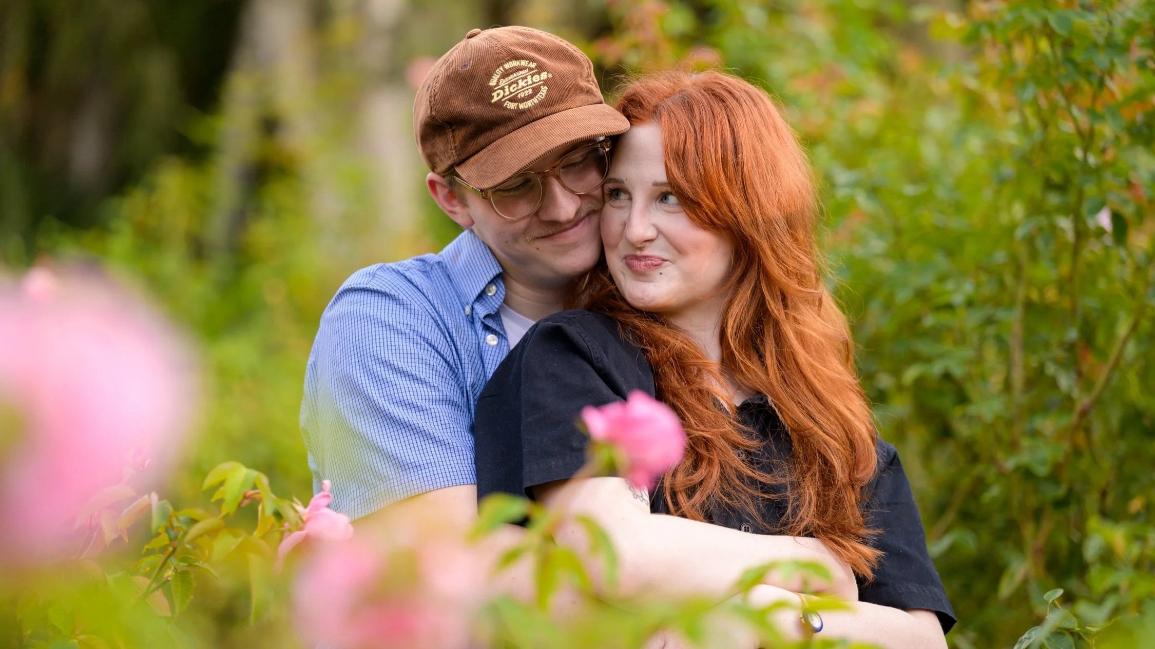 A young couple hugging outdoors among pink flowers and green foliage, smiling and looking happy.