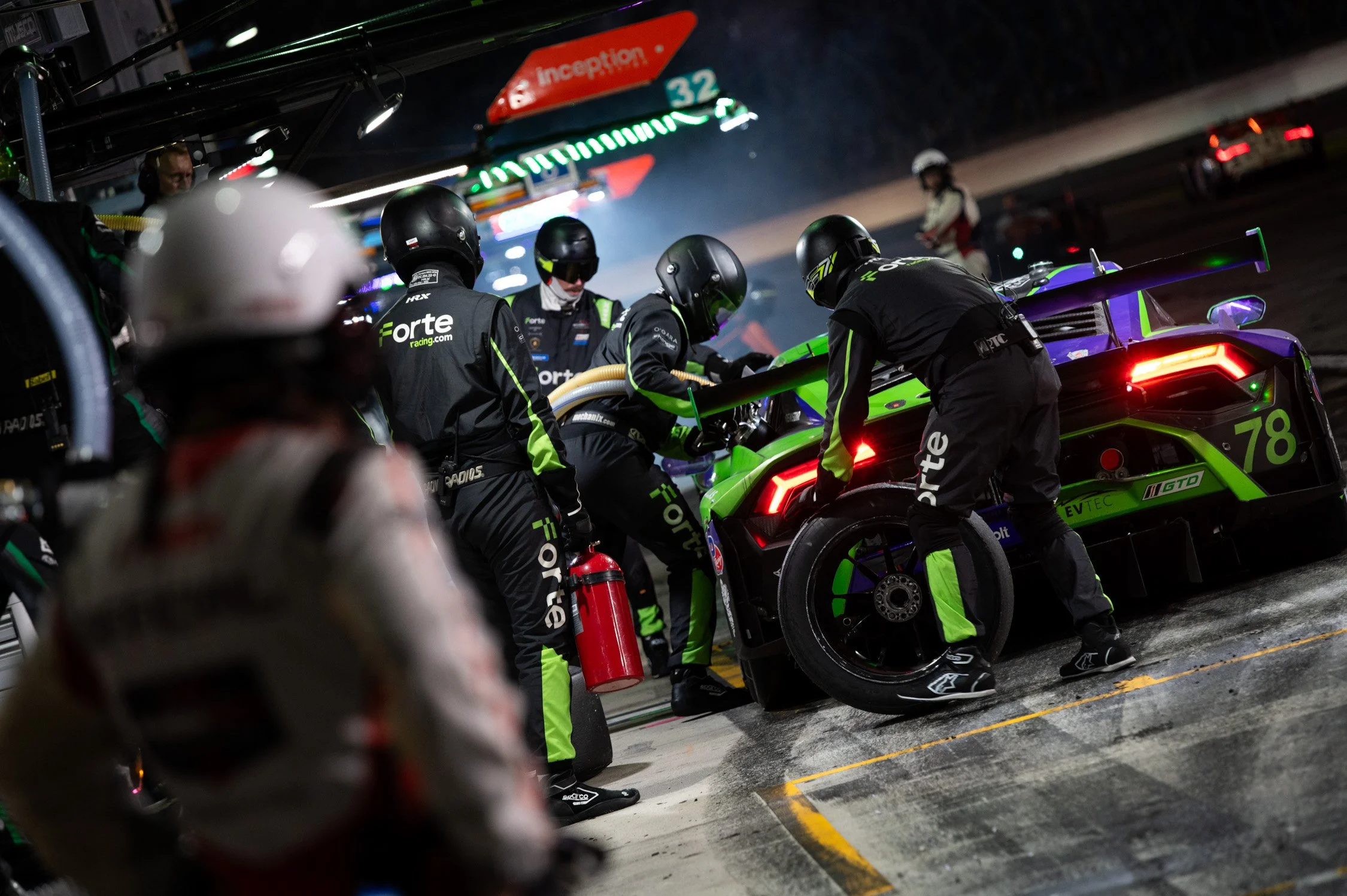 Race car pit crew working on a green and purple race car during a night race, with team members in black uniforms and helmets, and one crew member holding a tire.