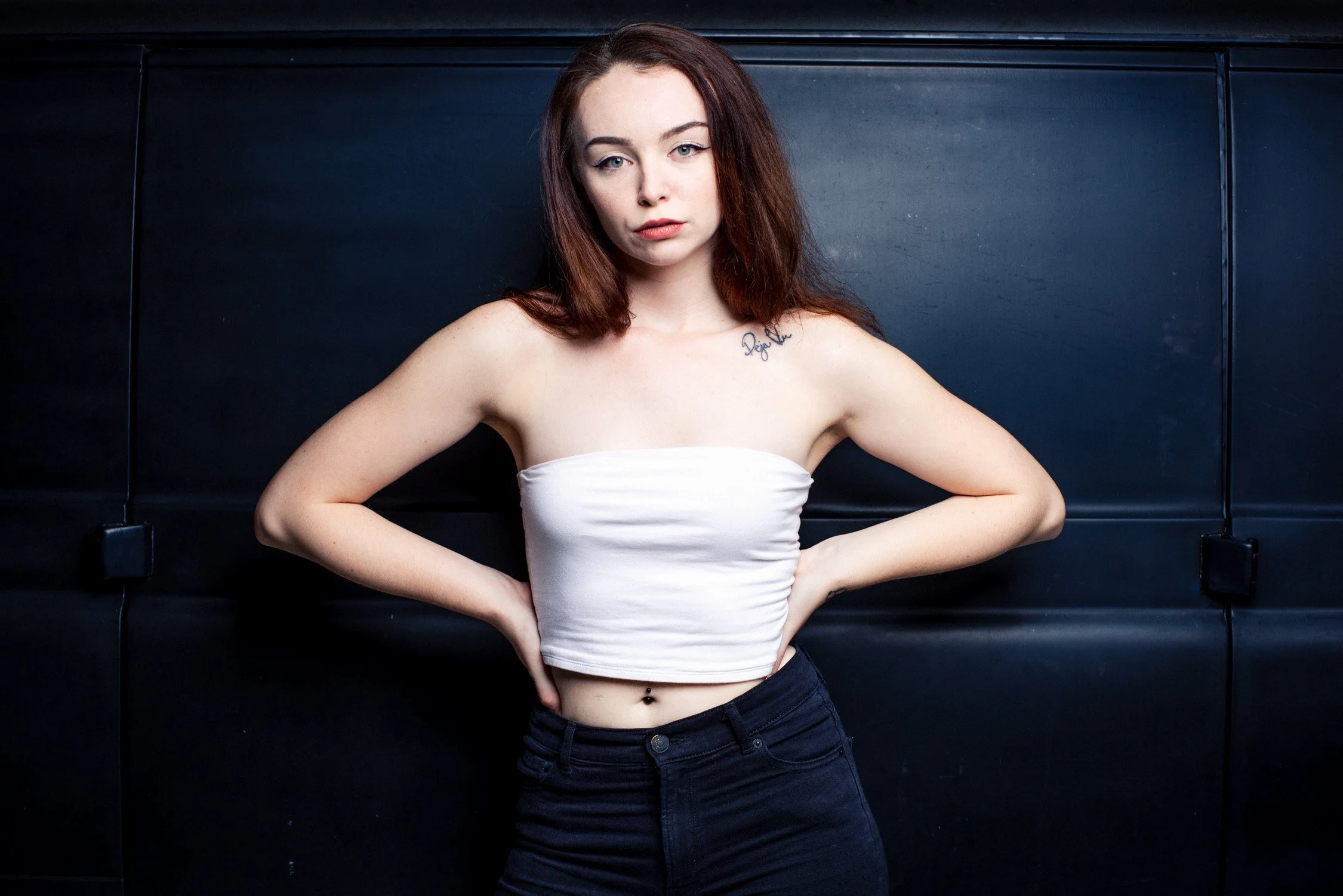 A young woman with red hair, wearing a white strapless top and black pants, standing against a dark background with her hands on her hips.