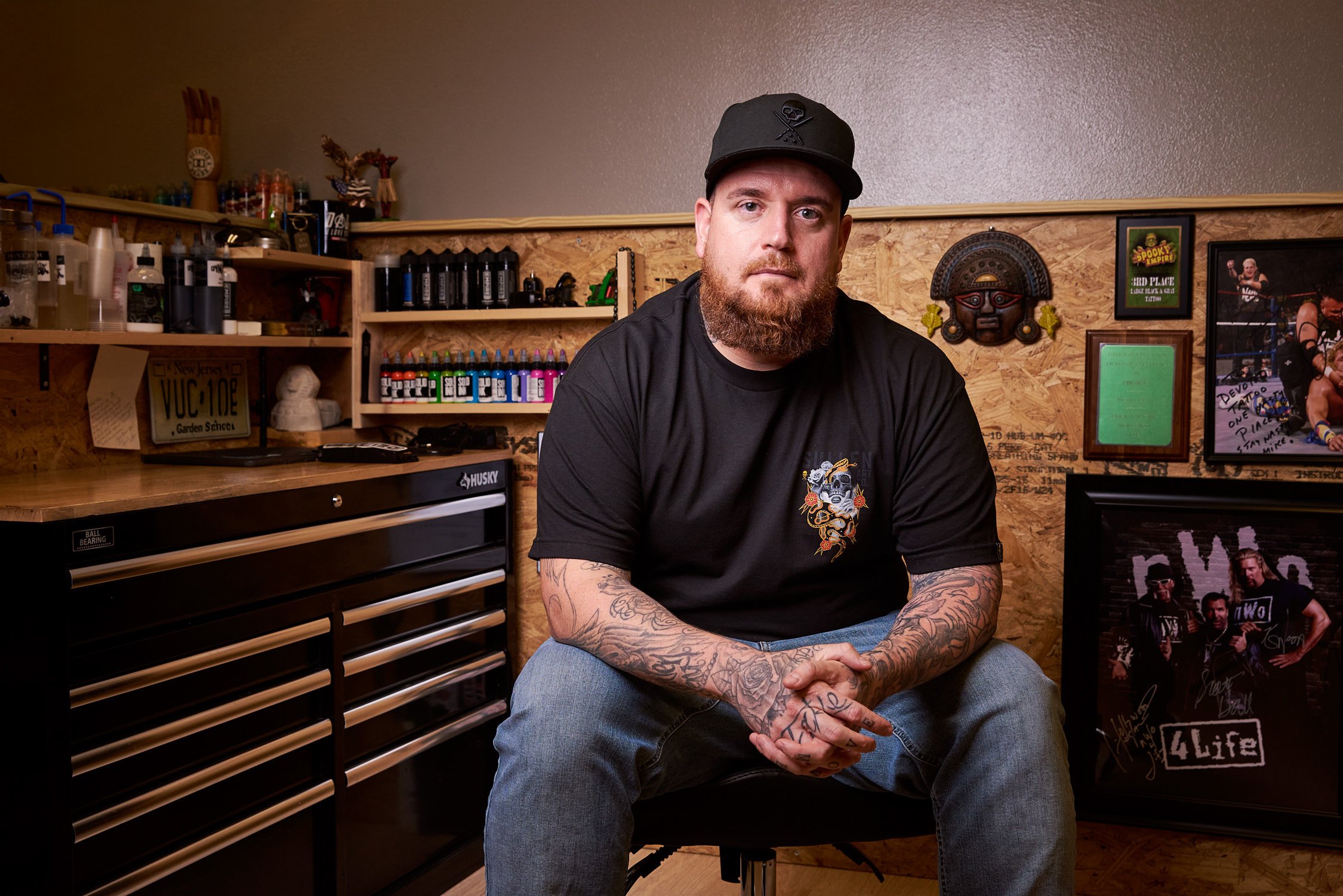 A man with a beard and tattoos wearing a black T-shirt and cap sitting in a workshop. The background features shelves with various tools and supplies, framed pictures, and artwork on the wall.