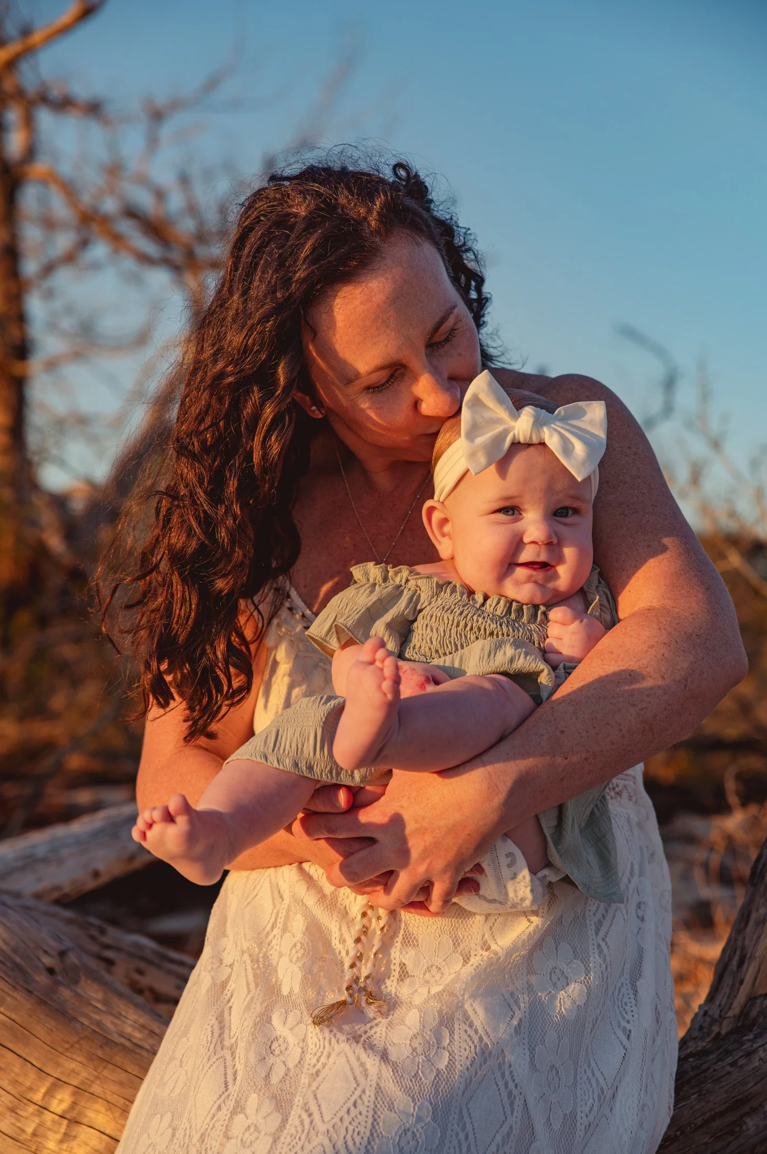 A woman with long brown curly hair holding a cheerful baby girl with a white bow on her head outdoors during sunset.