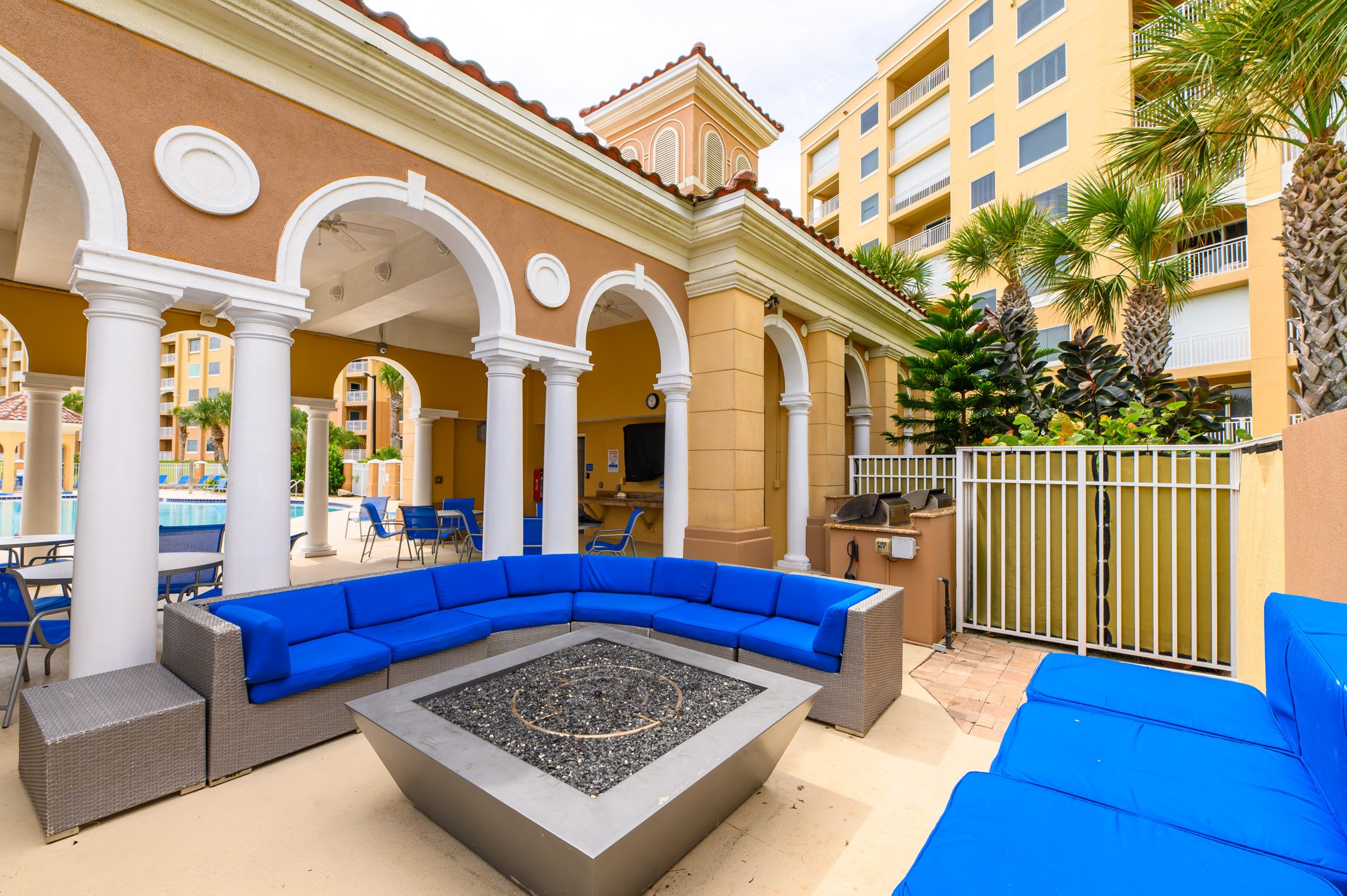 Outdoor lounge area with blue cushioned seating around a fire pit, surrounded by palm trees, and a pool in the background. Beige and yellow buildings with arches and columns are also visible.