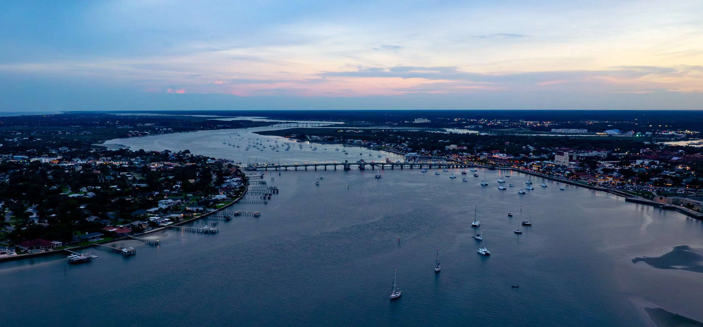 An aerial view of a coastal city at dusk with a river, boats, a bridge, and a cityscape in the distance.
