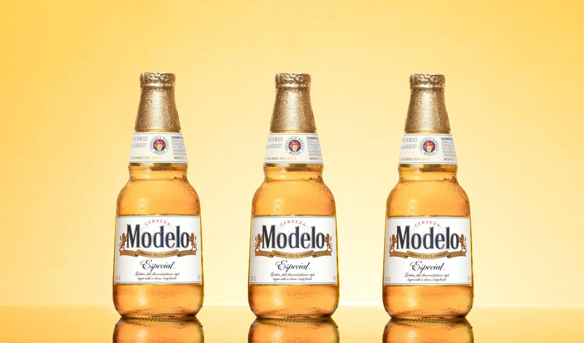 Three bottles of Modelo Especial beer on a reflective surface against a yellow background.