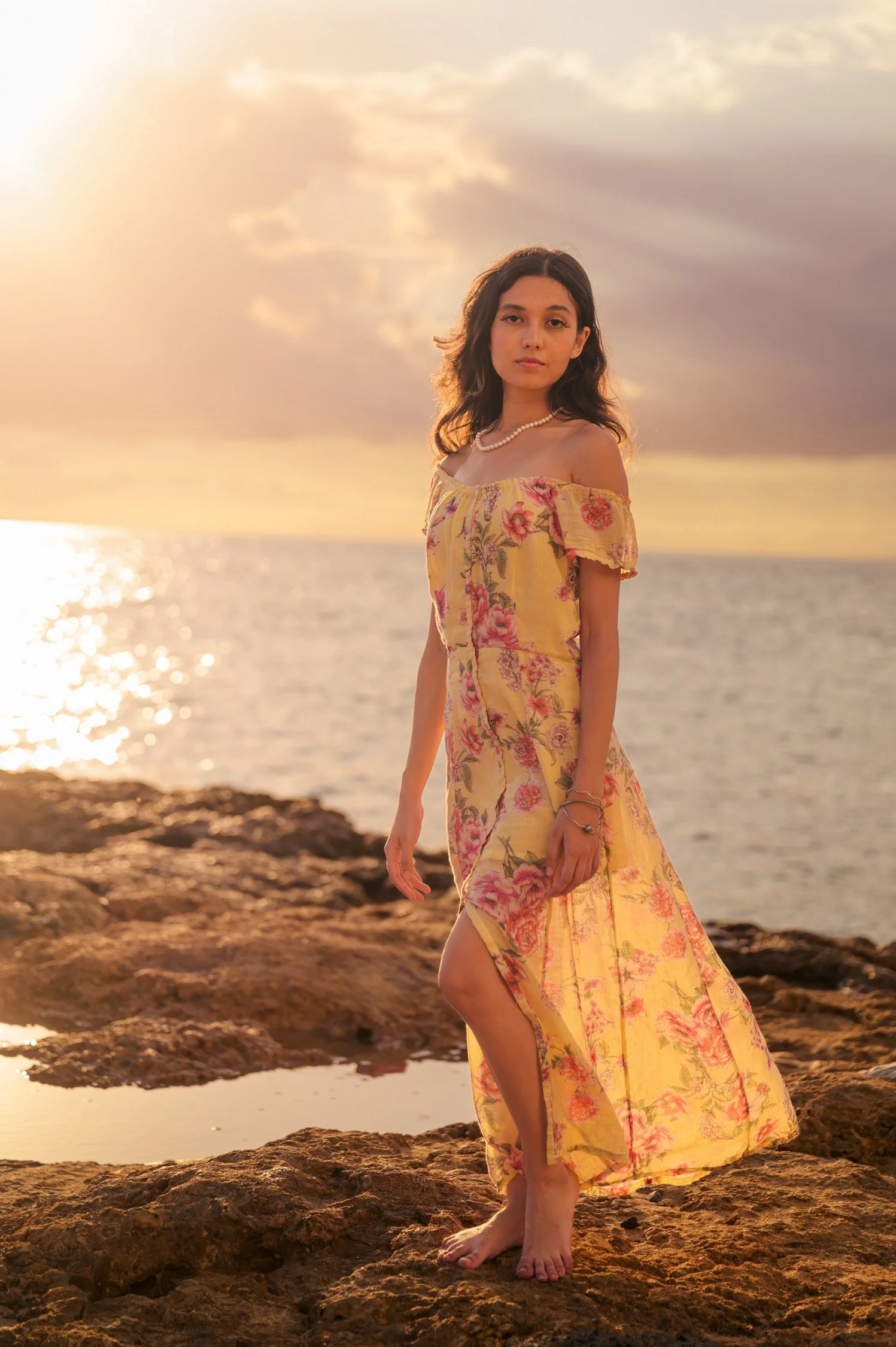 A young woman in a yellow floral dress standing barefoot on rocks by the ocean during sunset, looking at the camera.