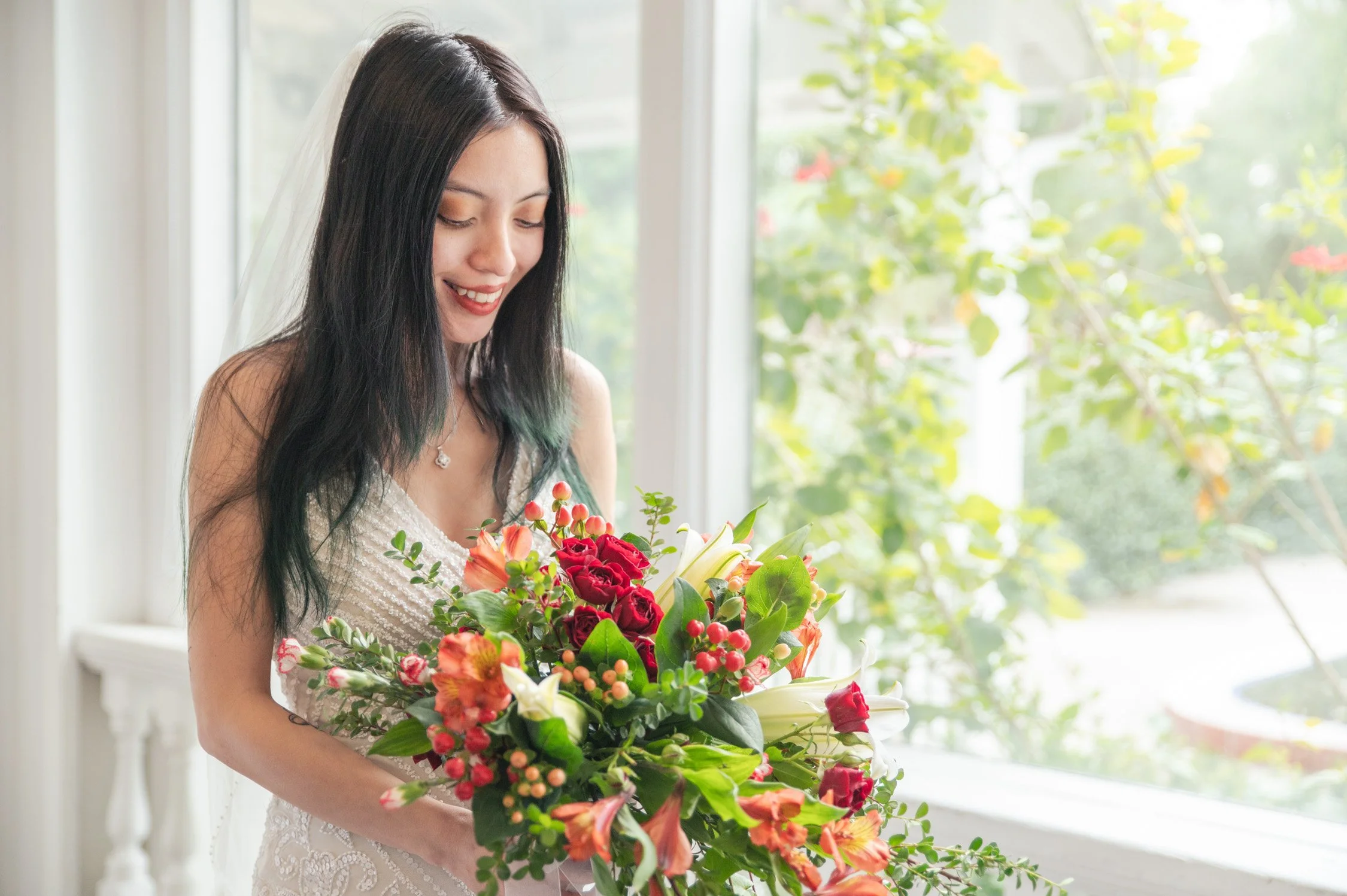 A woman in a wedding dress smiling while holding a bouquet of red, white, and orange flowers near a window with greenery outside.