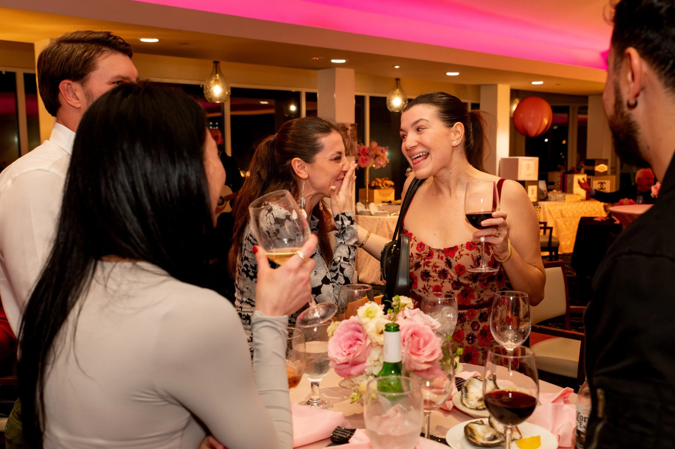 A group of people gathered around a table at a celebration, chatting and laughing, with drinks and flowers on the table, in a warmly lit venue.