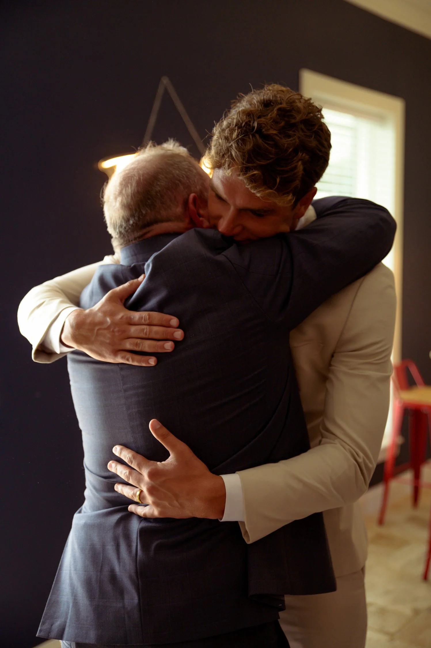 Two men in suits hugging warmly inside a room with a dark wall and a window in the background.