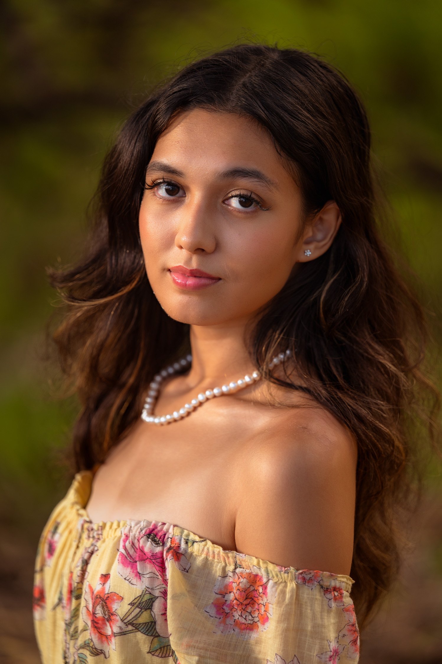 Portrait of a young woman with long, wavy hair, wearing a floral off-shoulder top, a pearl necklace, and earrings, outdoors with a blurred green background.