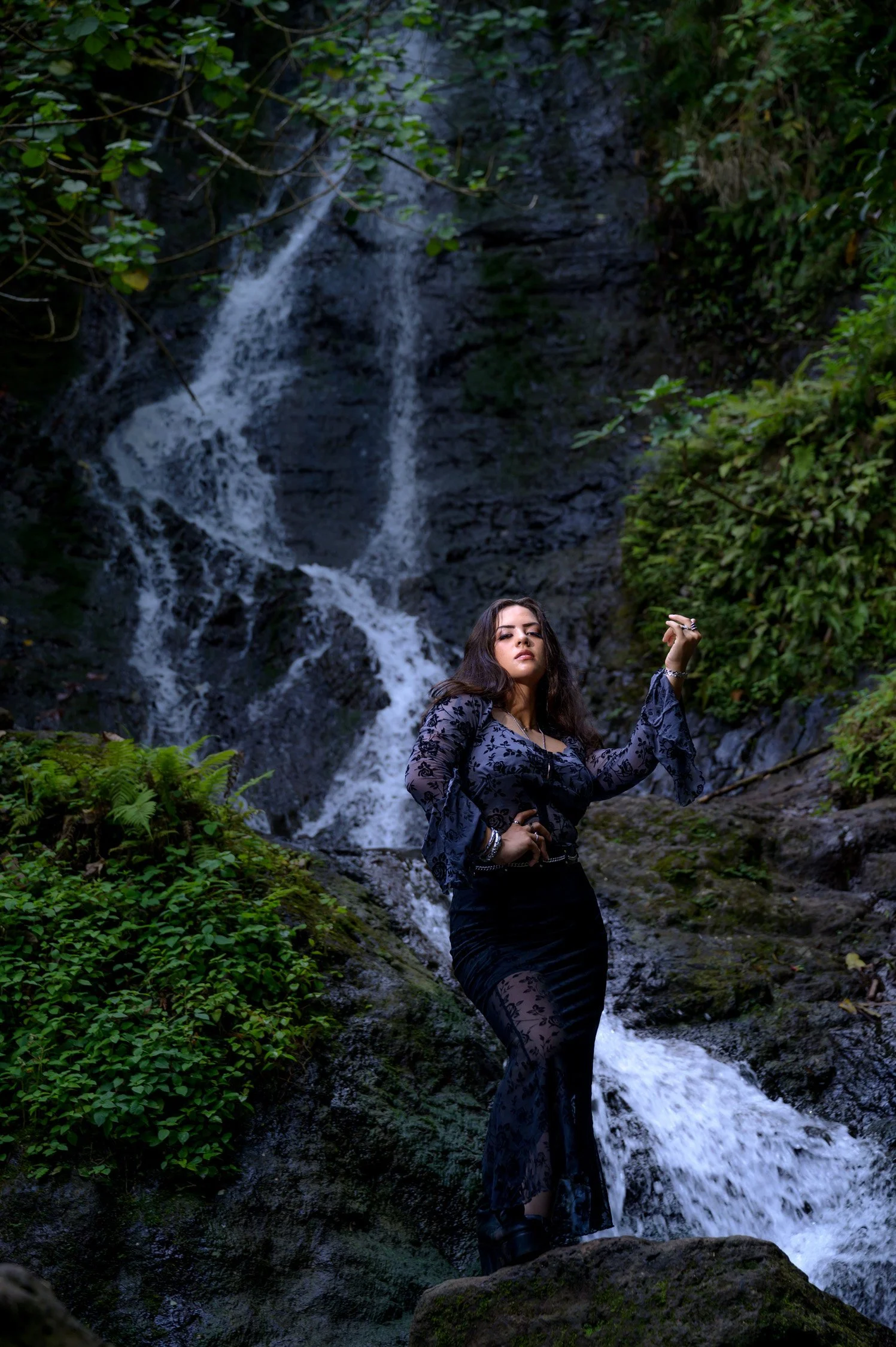 A woman in a black lace dress standing on rocks in front of a waterfall surrounded by greenery.