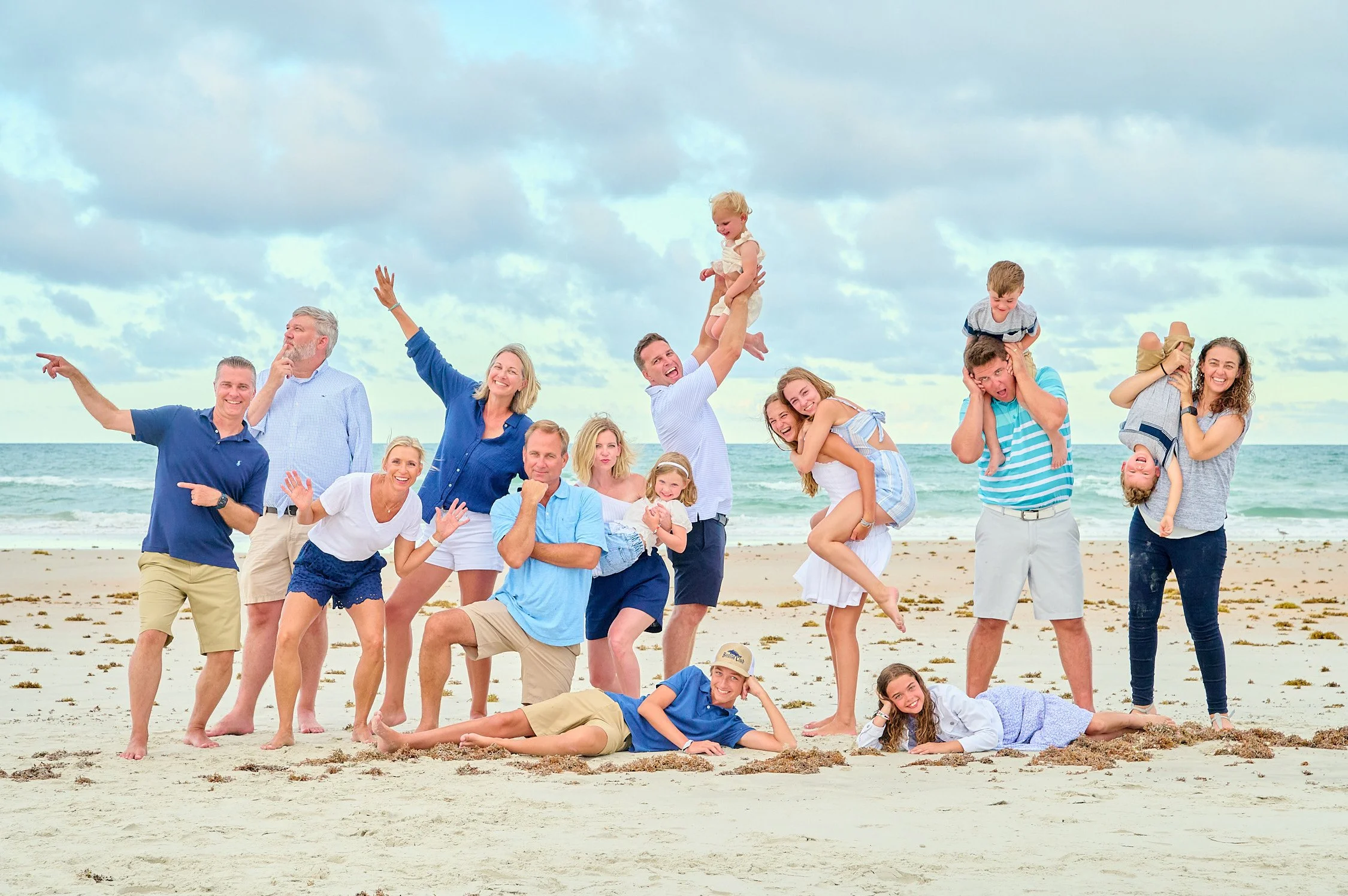 A large group of people, including children and adults, enjoying a day at the beach. Some are standing, some are sitting or lying on the sand, and a few are playing or posing for the photo. The sky is partly cloudy, and the ocean is visible in the ba