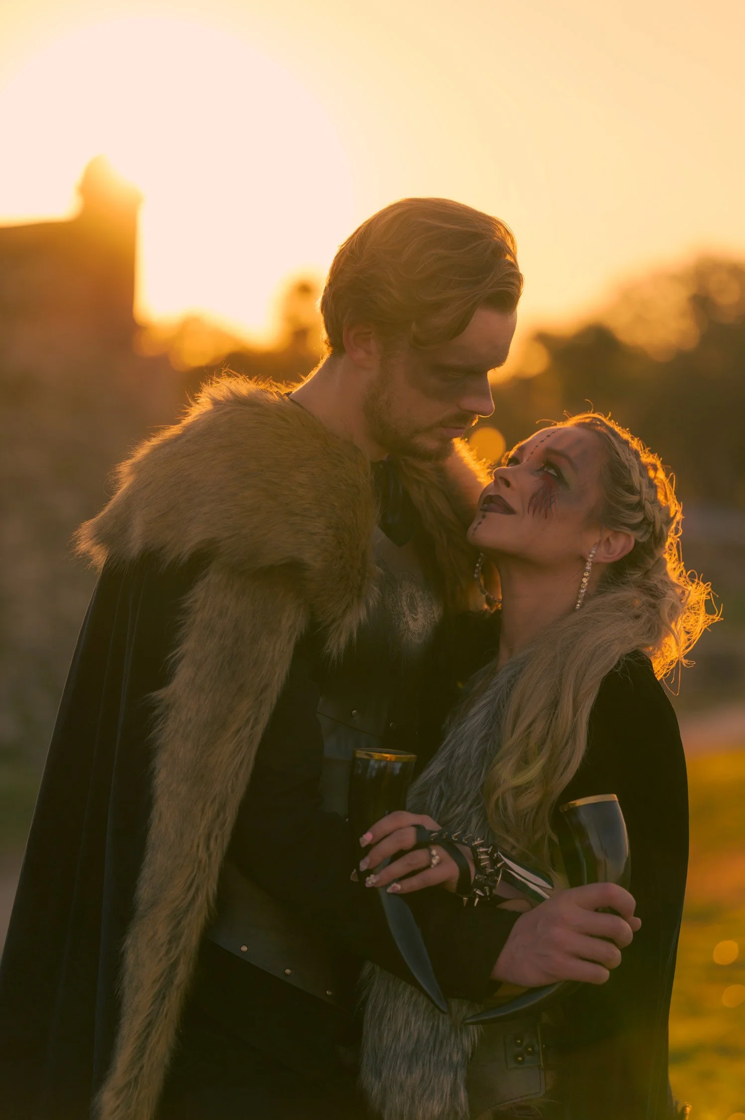 Two people dressed in fantasy costumes, one with tribal makeup, holding wine glasses at sunset.