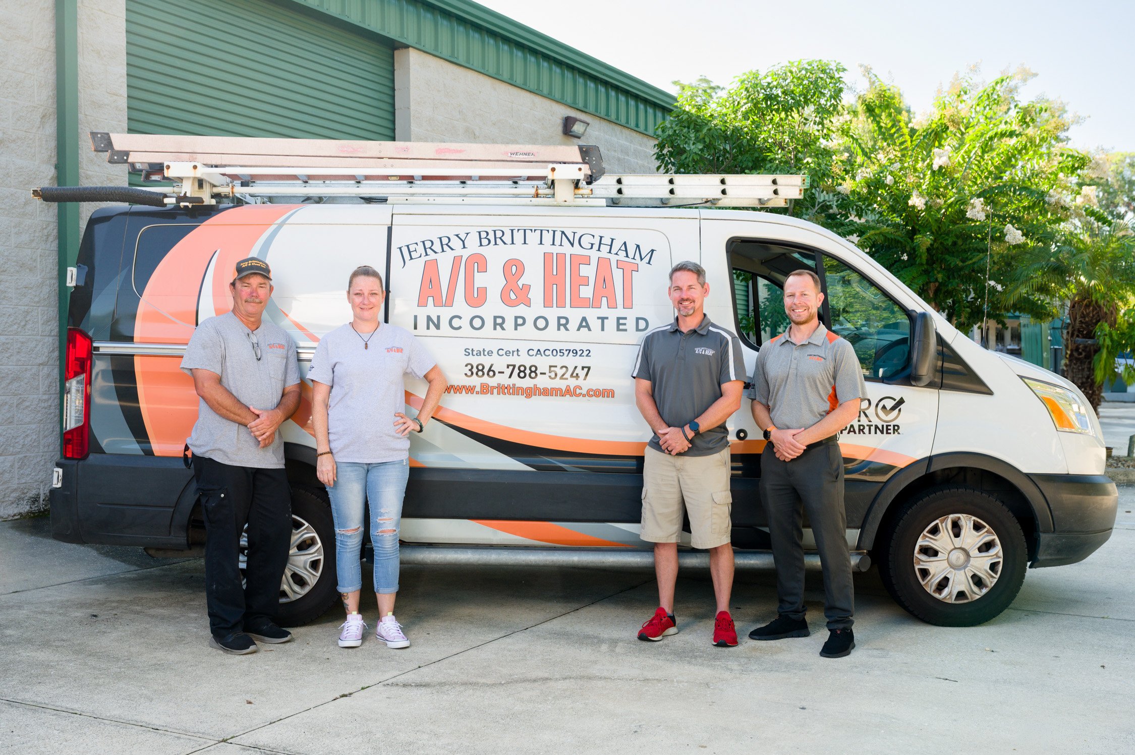 Four people standing in front of a company van with the logo 'Jerry Brittingham A/C & Heat Incorporated,' parked outside a building with trees and a green garage door.