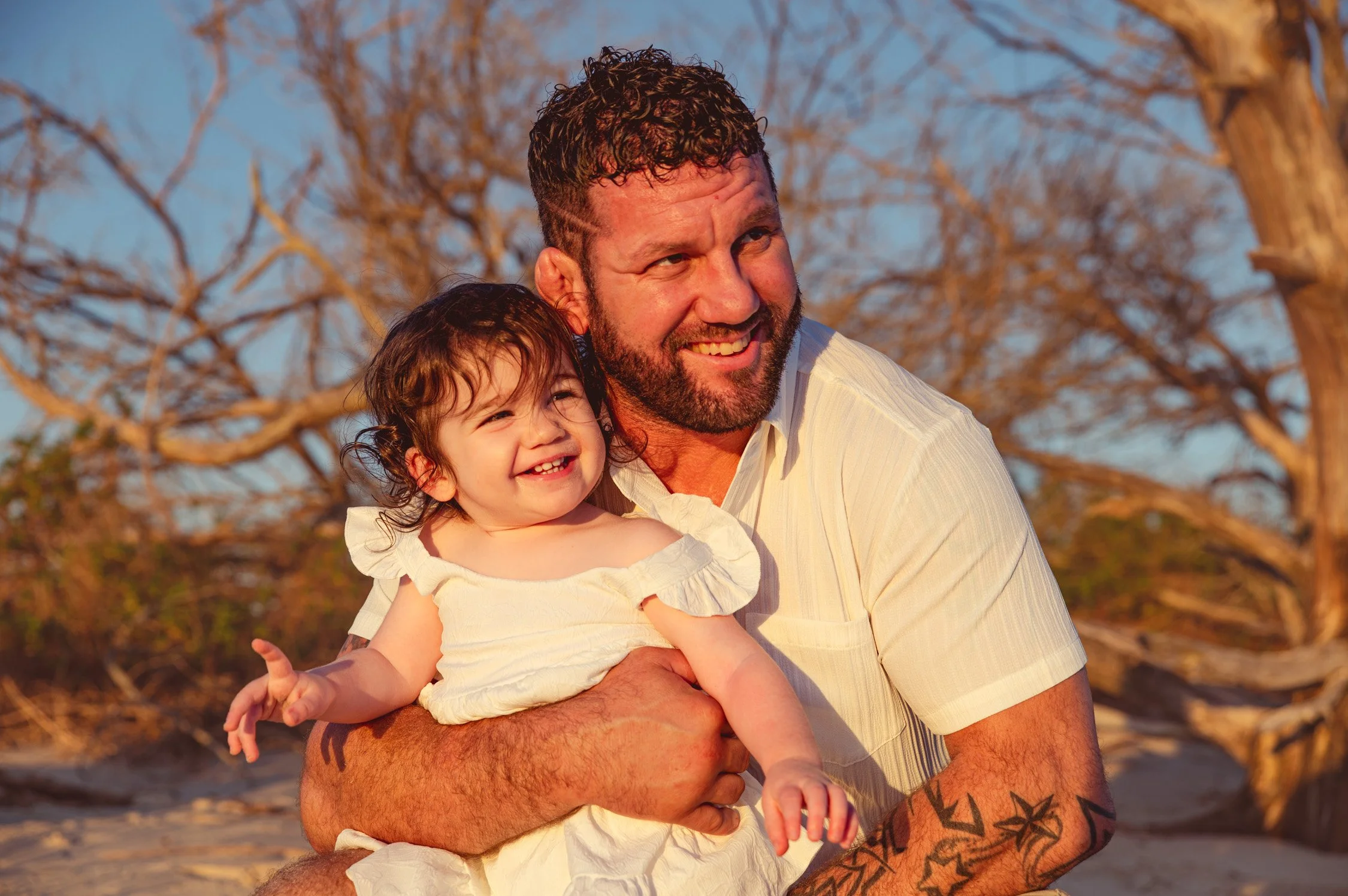 A man holding a young girl outdoors during sunset, smiling, with leafless trees in the background.