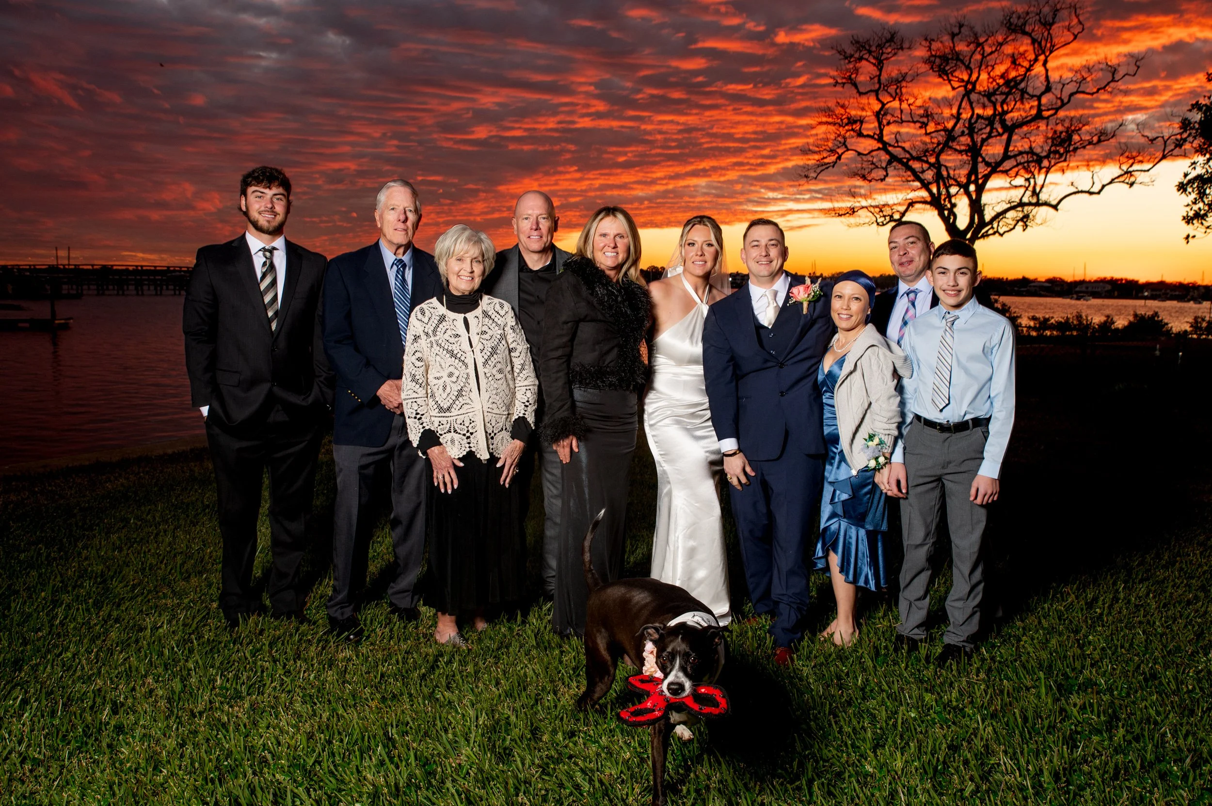 A group of people dressed in formal attire, including a bride in a white wedding dress and a groom in a navy suit, standing together outdoors during sunset with a colorful sky and lake in the background, along with a dog holding a red toy