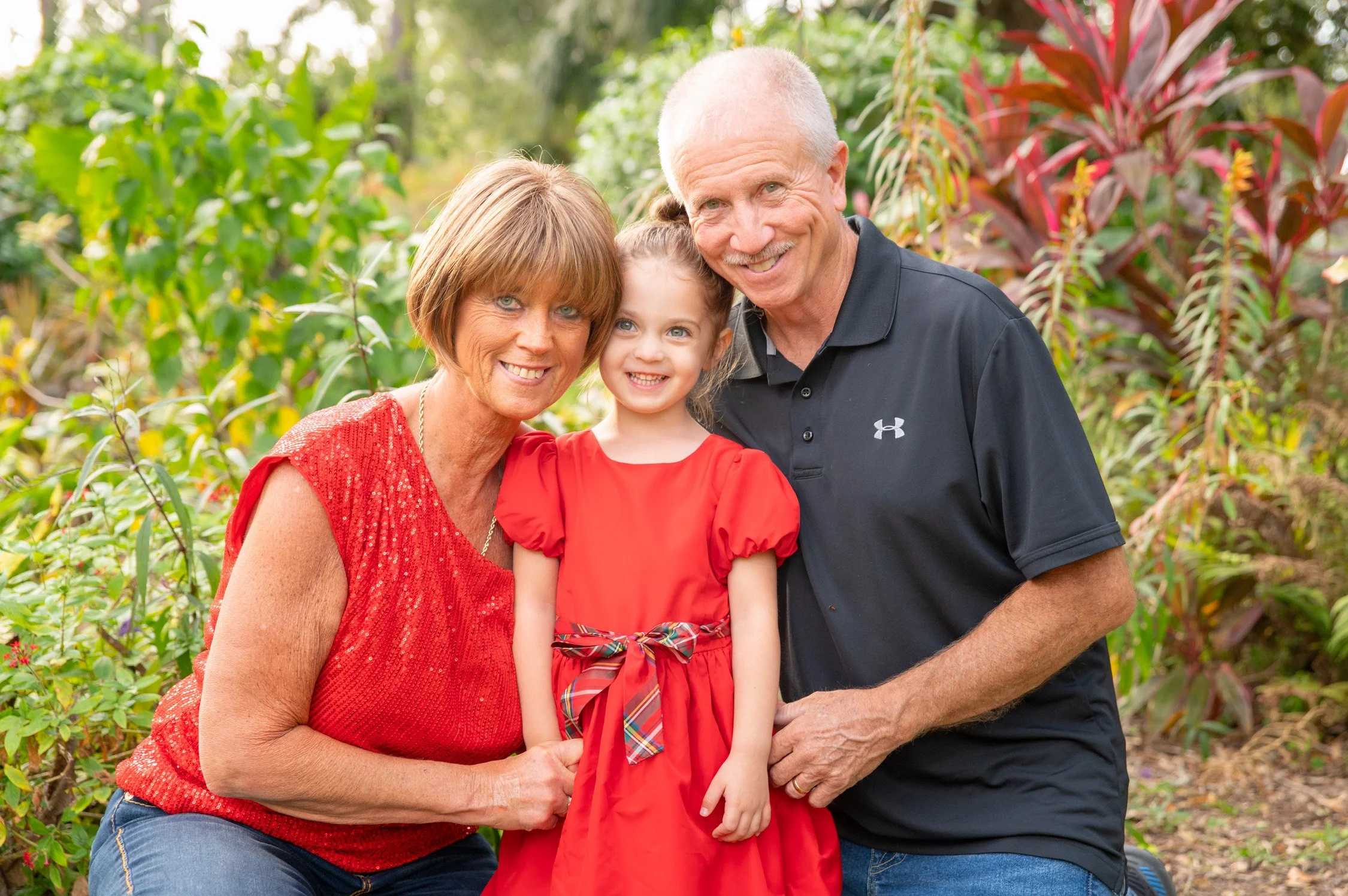 A happy family of three outdoors, with a woman, a little girl, and a man, all smiling and dressed in red and black, surrounded by lush green plants and trees.