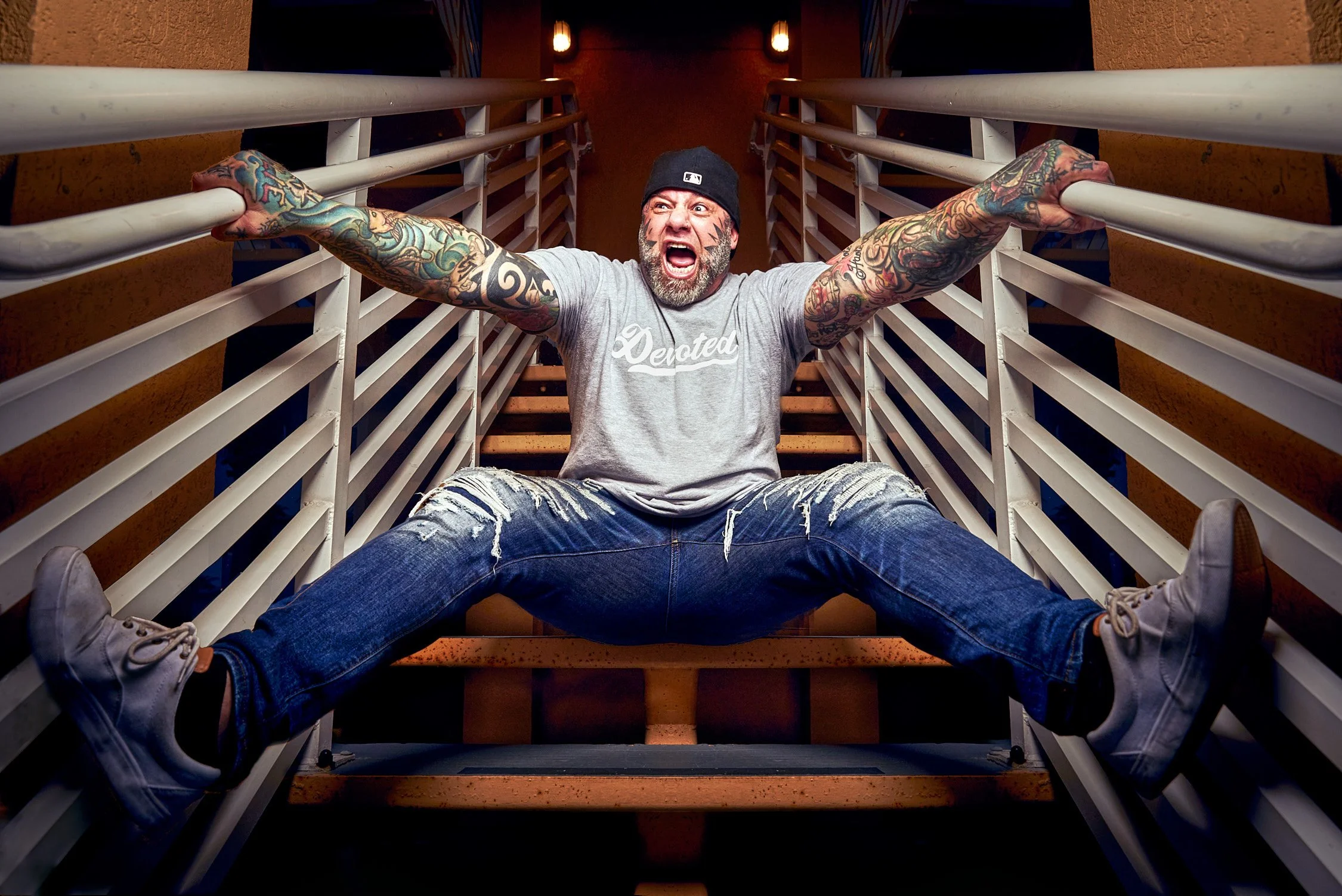 A man with colorful tattoos on his arms, wearing a gray t-shirt, ripped jeans, and a black beanie, is sitting with legs spread on stairs, holding onto railings, yelling or expressing excitement in a dramatic pose.