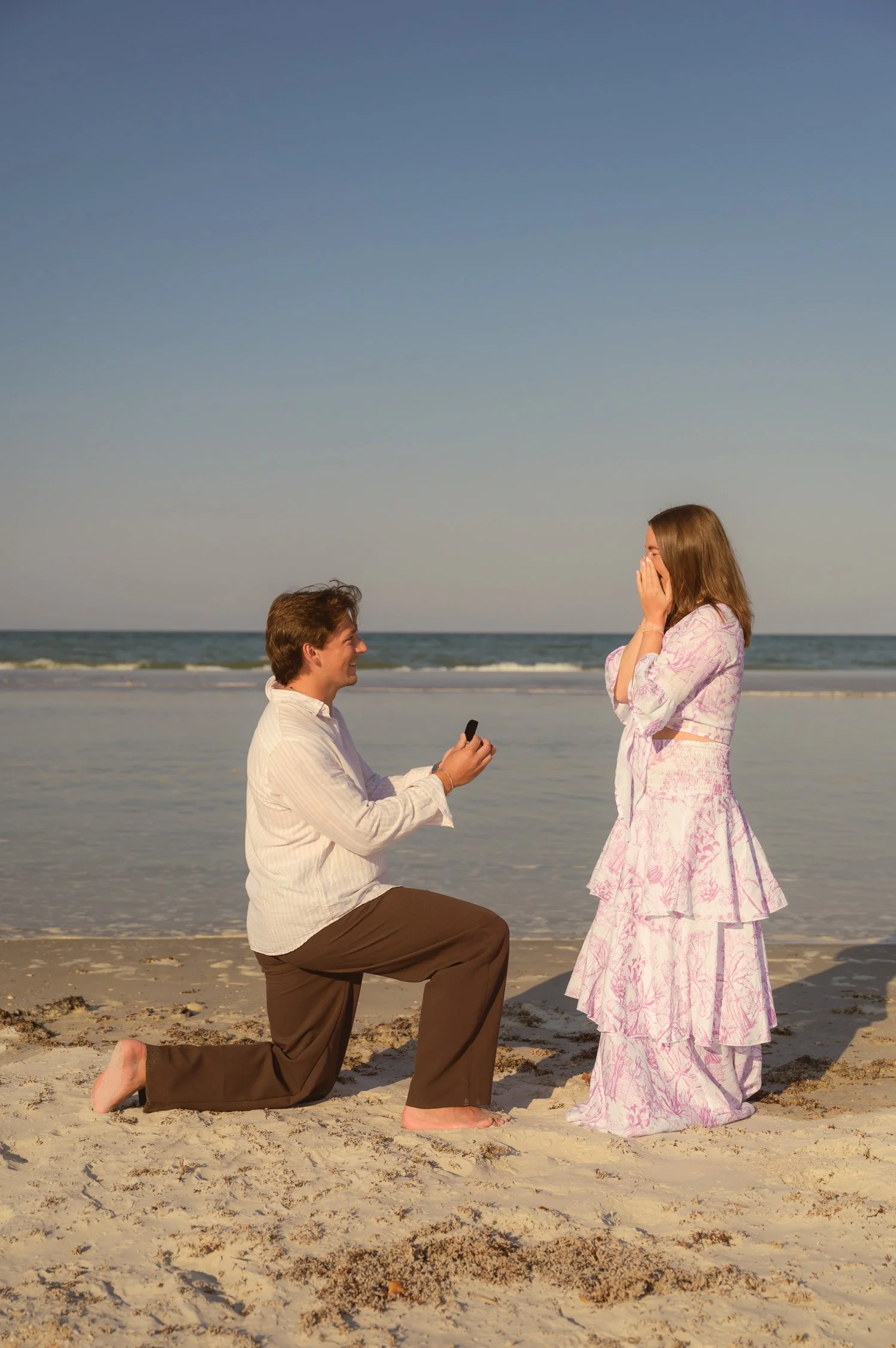 A man is kneeling on the sand, proposing to a woman on the beach. The woman is covering her face with her hands, appearing surprised and happy. The scene takes place near the ocean with a clear sky.