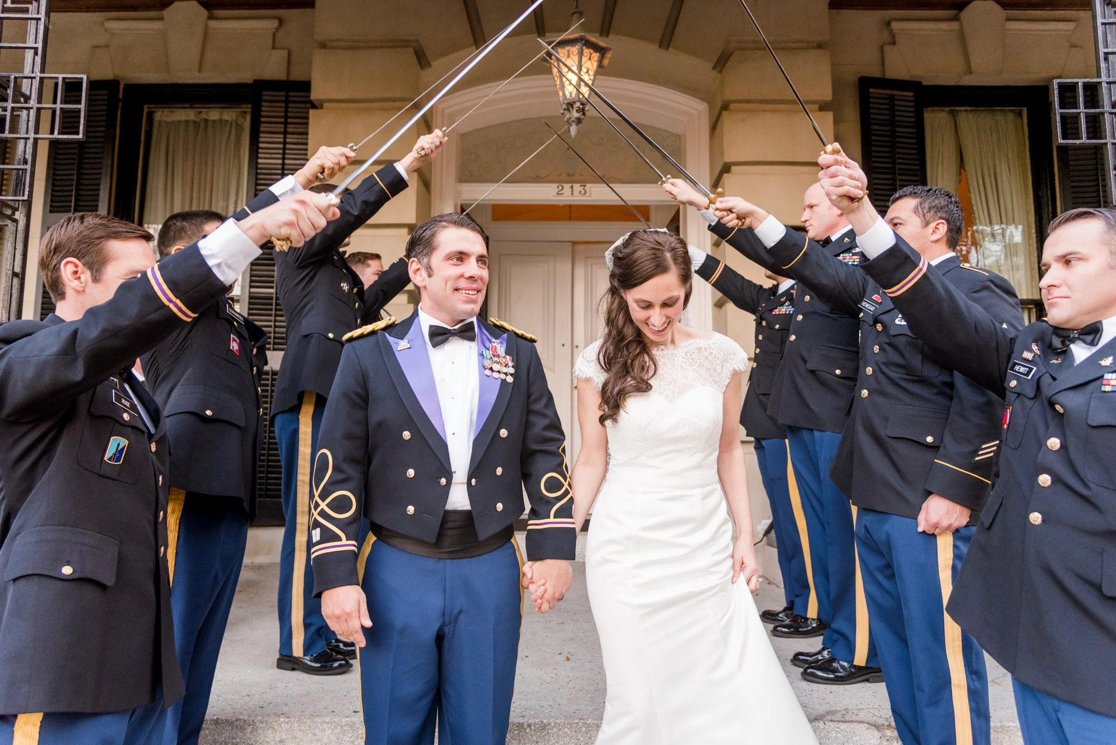 A bride and groom holding hands and smiling as soldiers in uniform form an arch with swords over their heads at their wedding outside a building.