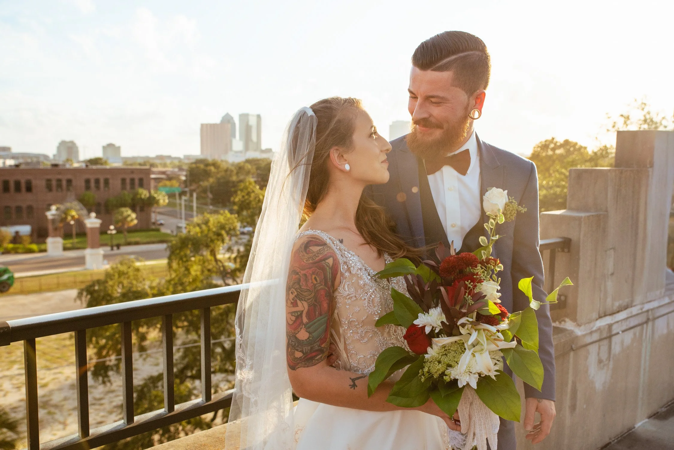 A bride and groom on a balcony during sunset, looking into each other's eyes. The bride holds a bouquet of flowers, and the groom wears a tuxedo with a black bow tie. The city skyline is visible in the background.
