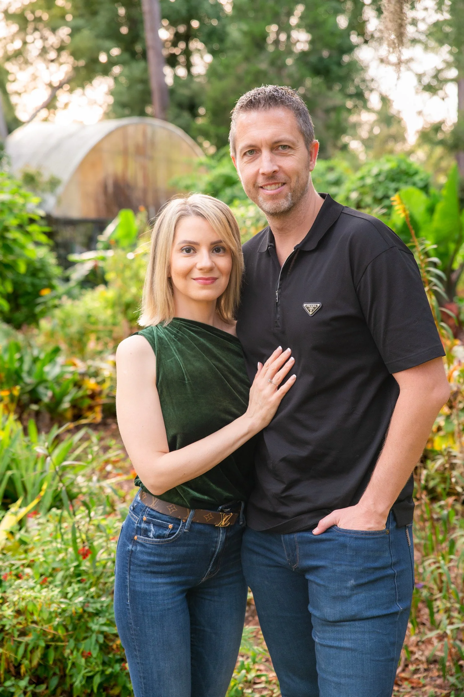 A man and woman standing close together outdoors with lush green foliage and a greenhouse in the background.