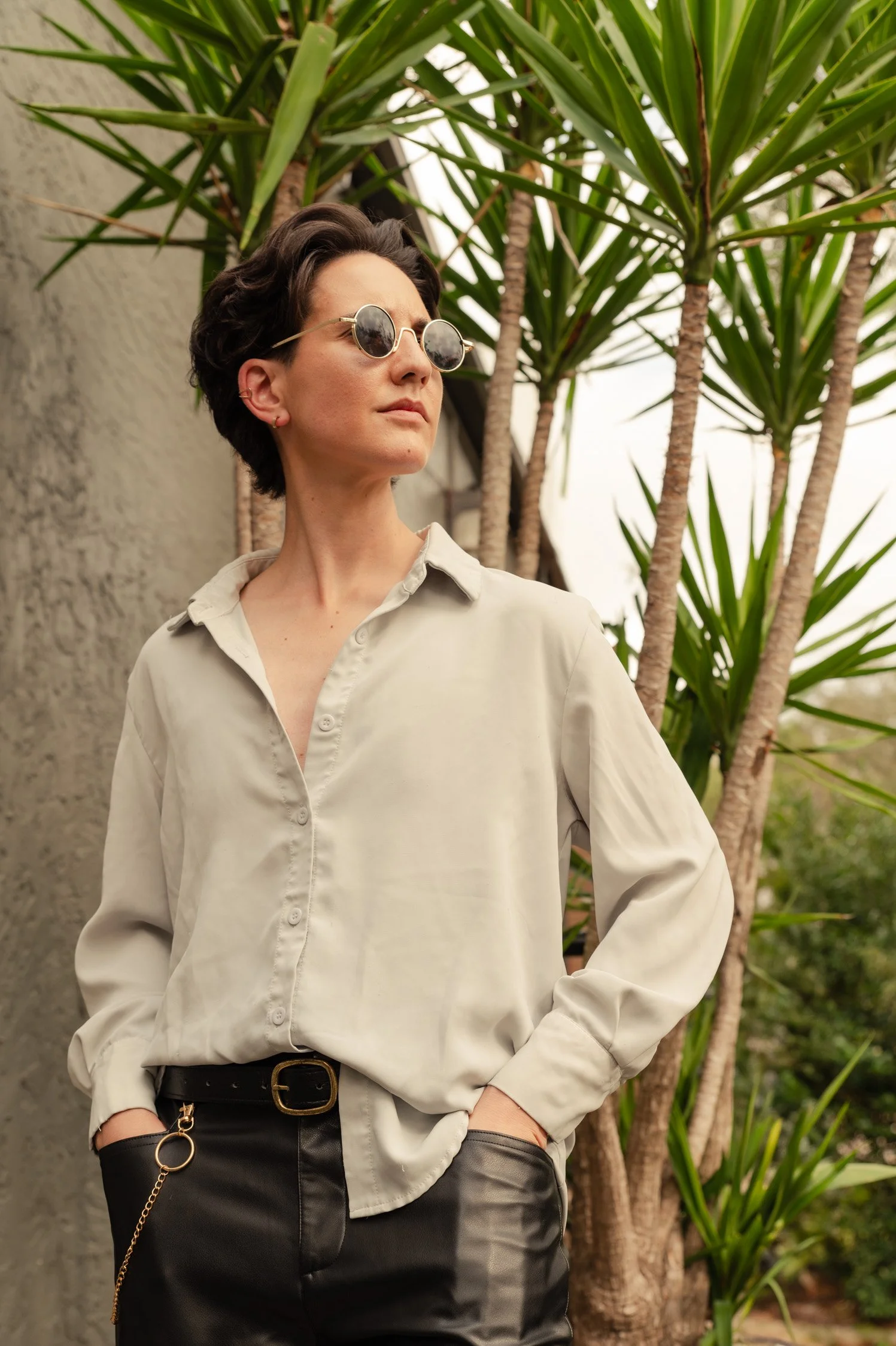 A woman with short dark hair wearing sunglasses, a cream-colored blouse, and black pants standing outdoors near tall green plants.