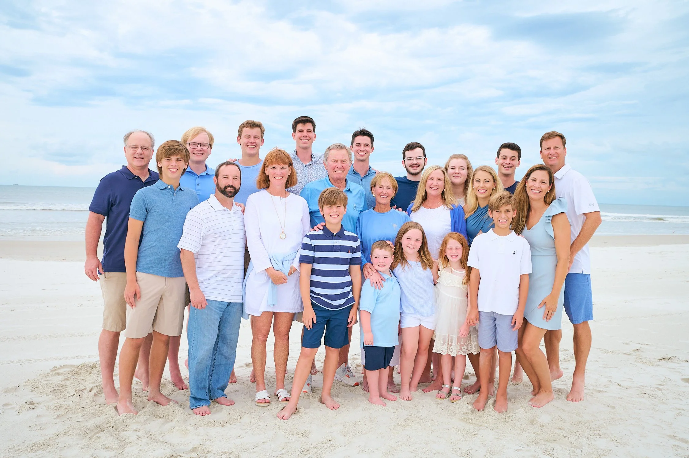 A large family of 20 members, including children and adults, posing together on a beach with sand and ocean in the background under a partly cloudy sky.