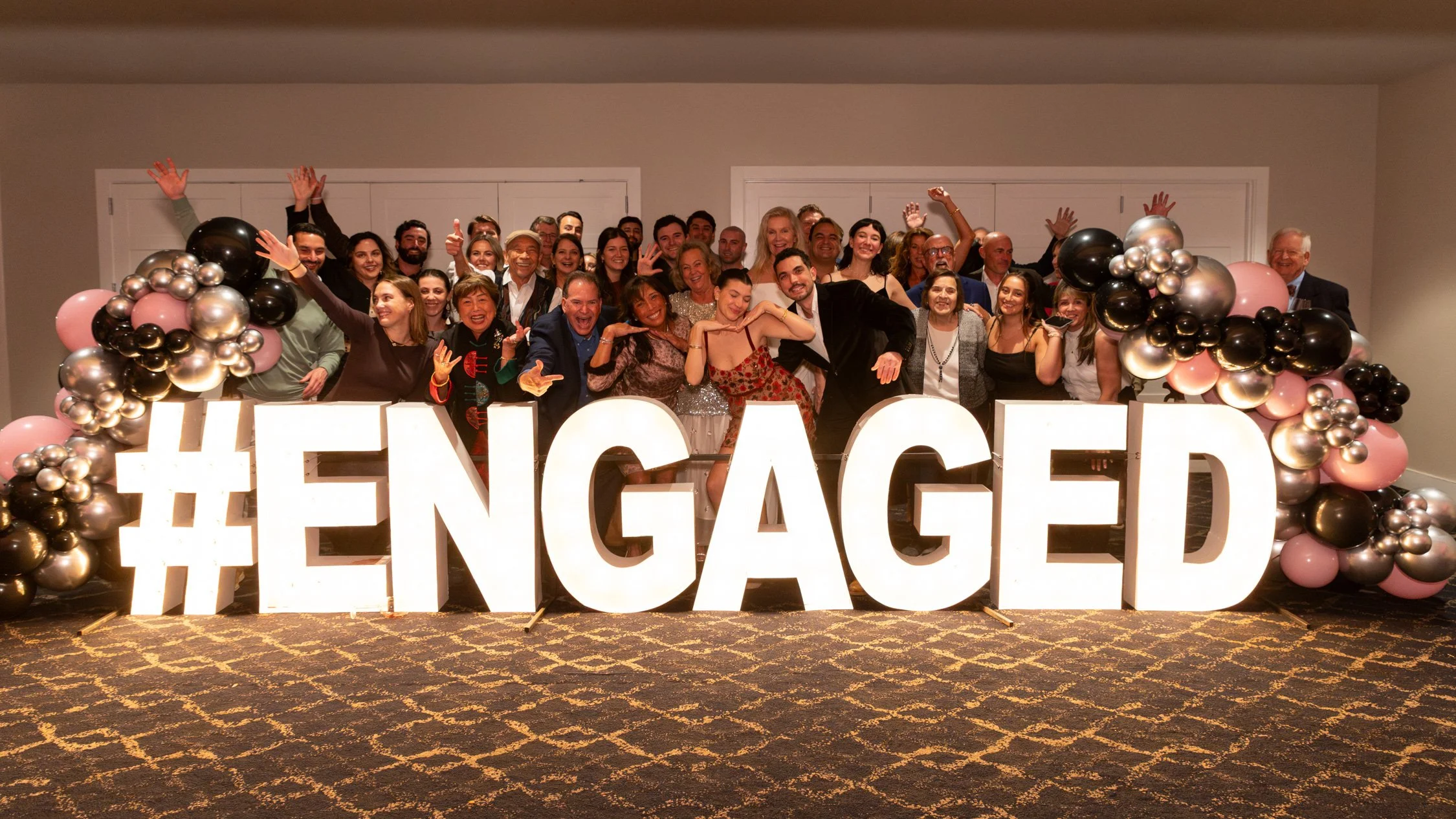 Group of people celebrating at an event, standing behind large illuminated letters spelling '#ENGAGED', with balloon decorations on each side.