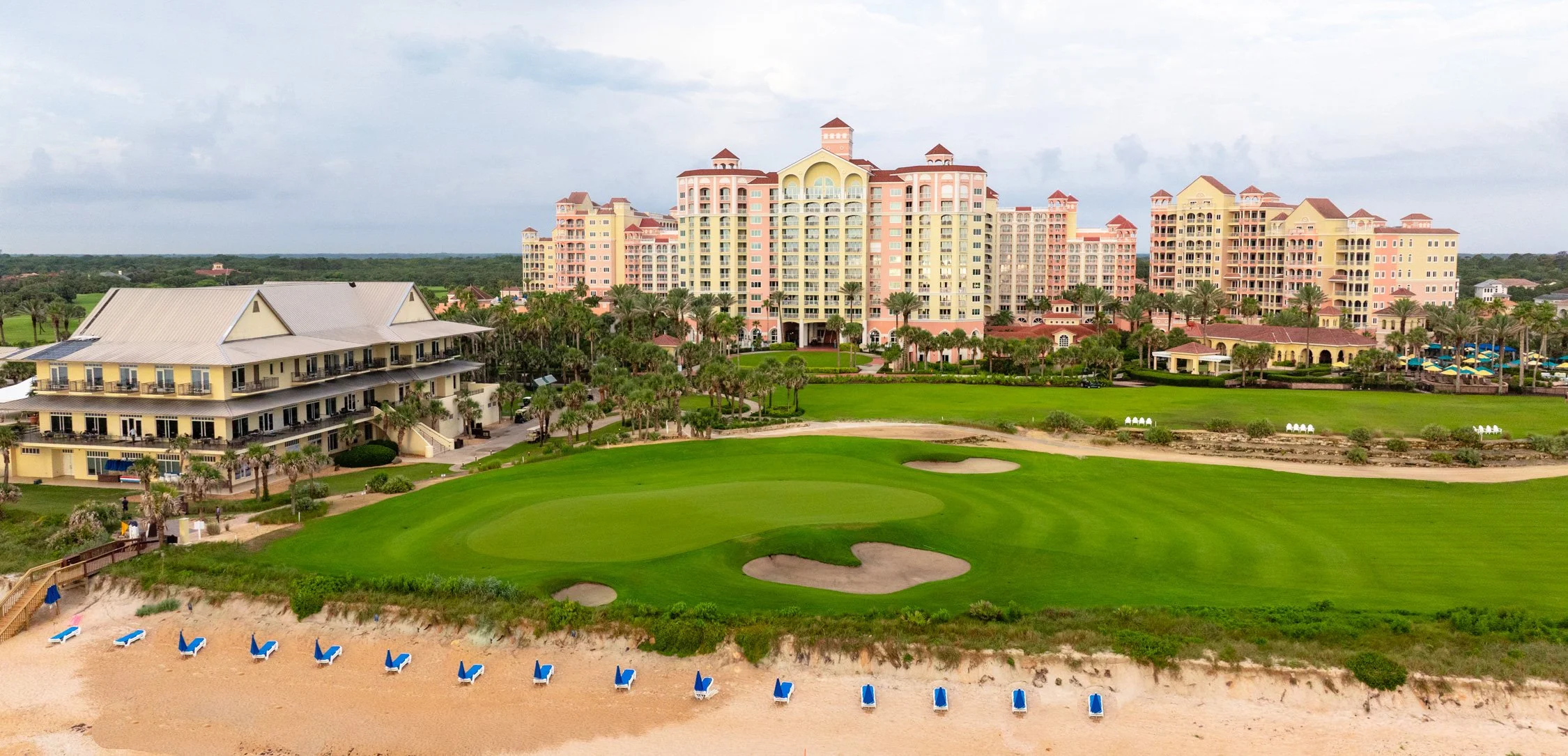 Beachside resort with golf course, sand bunkers, and colorful buildings in the background, under cloudy sky.