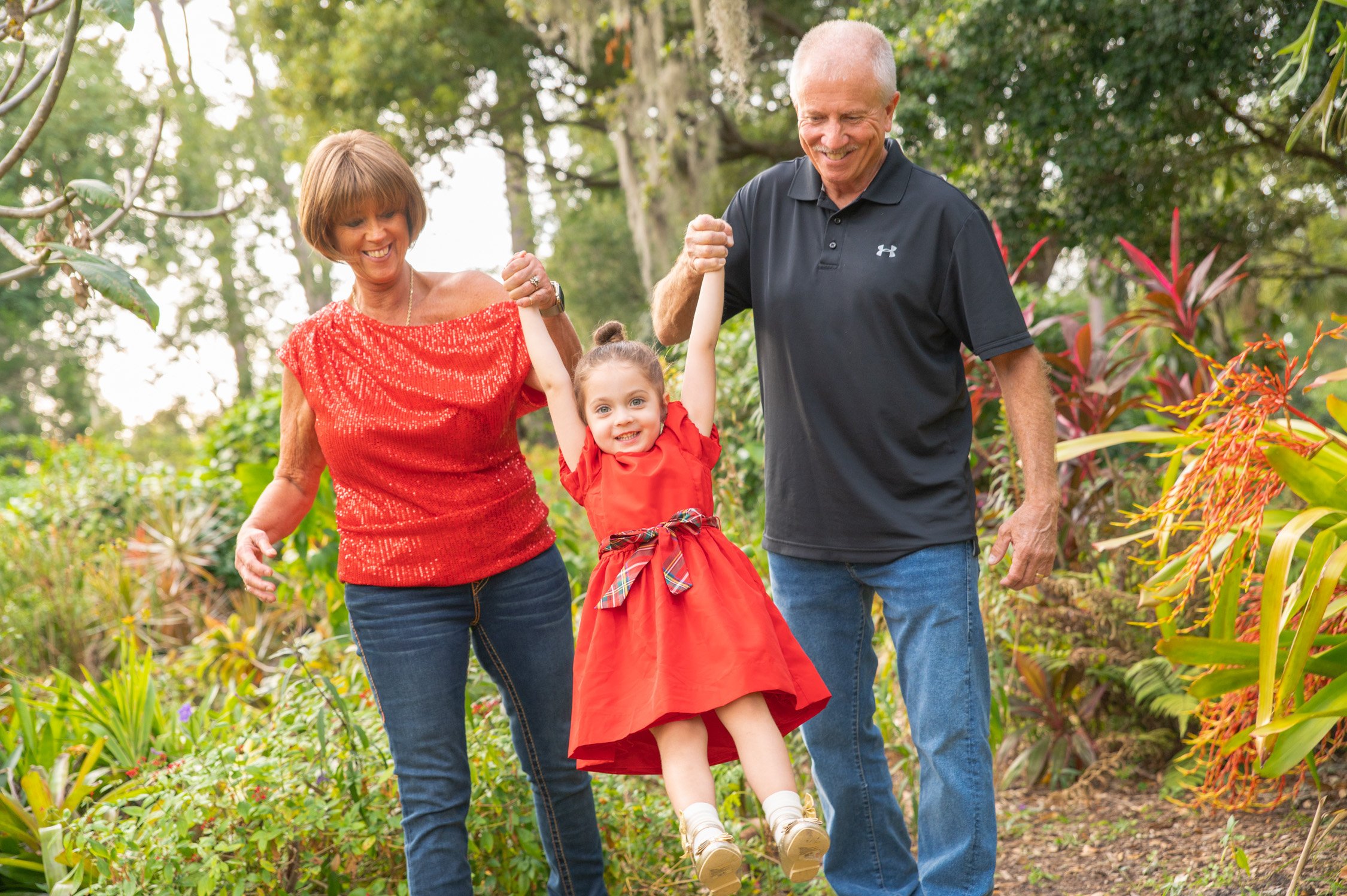 Family of three, two seniors and a young girl, walking in a garden. The young girl is being lifted by her grandparents, smiling and enjoying the moment.