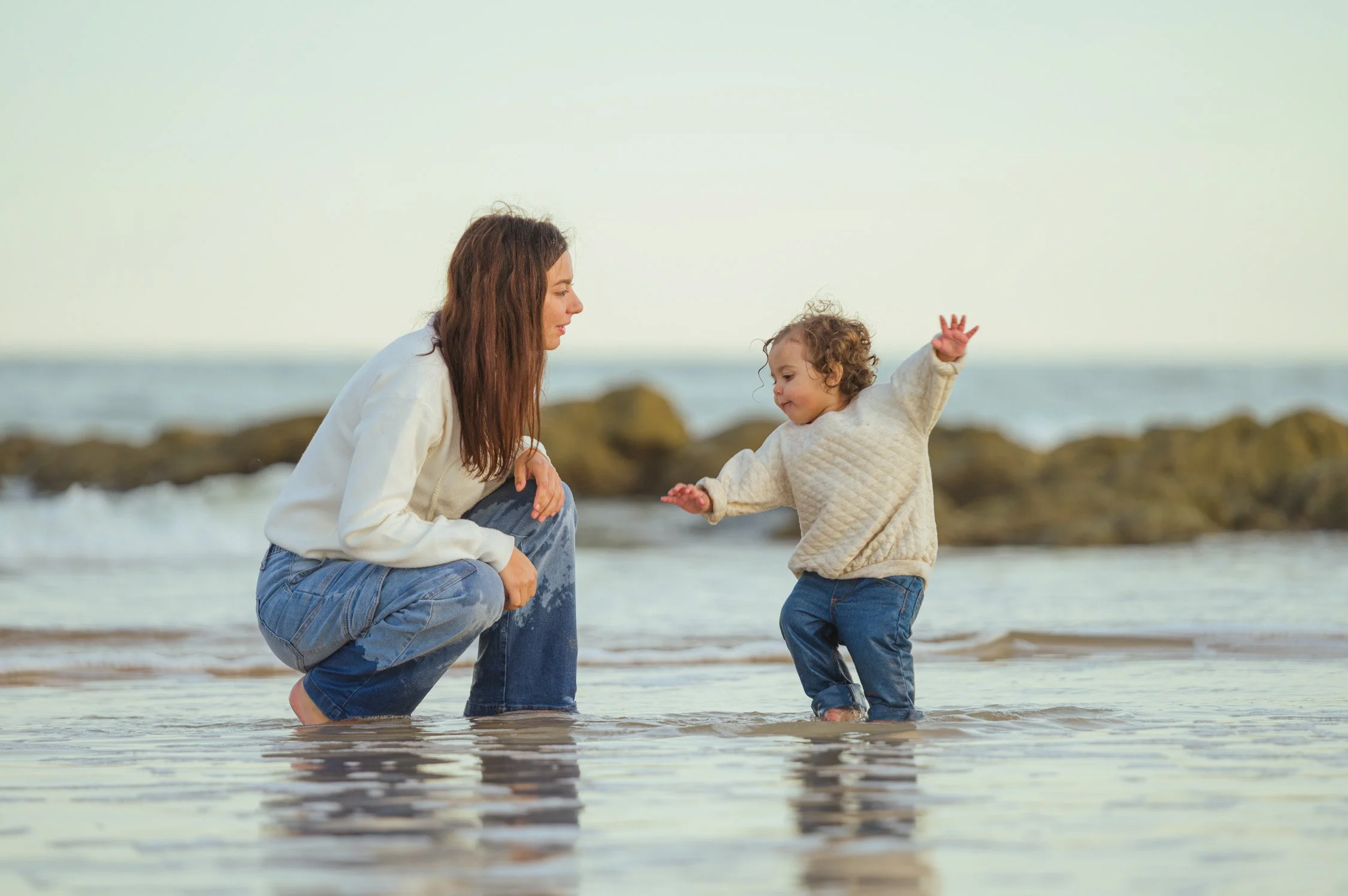 A woman and a young child are playing in shallow water at the beach, with rocks in the background.