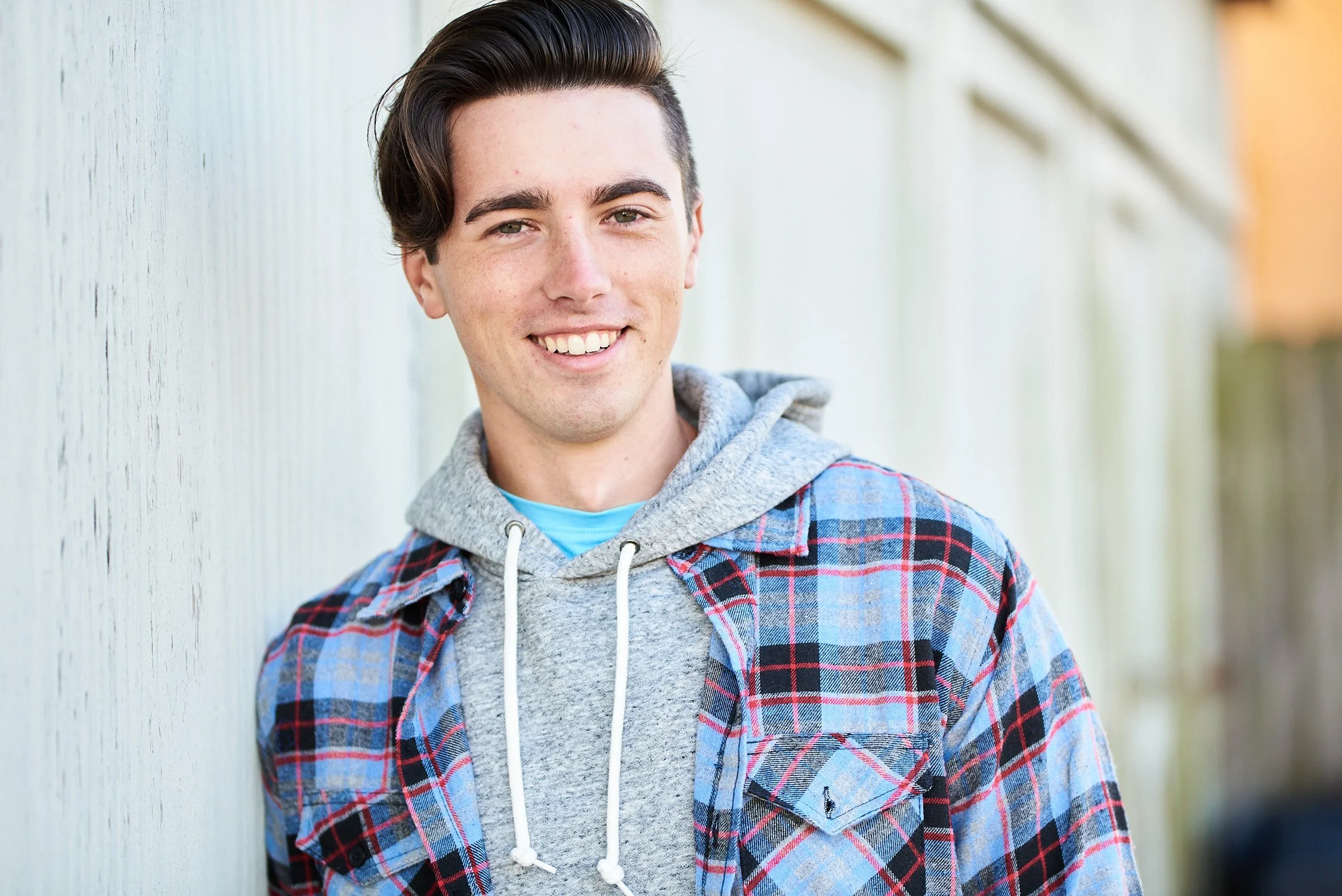 A young man with dark hair, wearing a gray hoodie under a red and blue plaid shirt, smiling and leaning against a white wooden fence outdoors.
