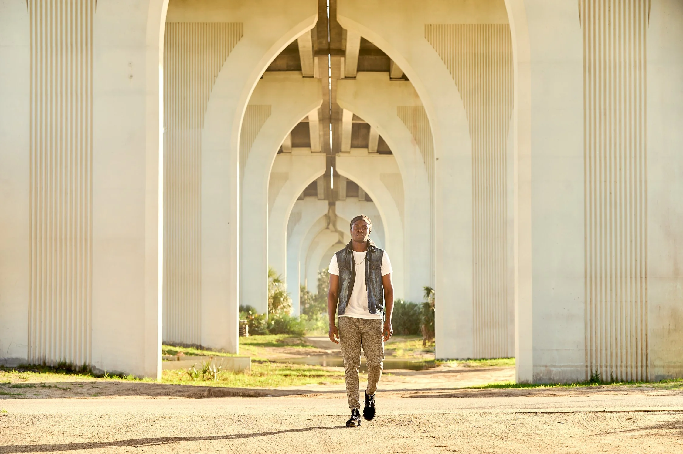 A young man walking under a large concrete bridge with arches during daytime.