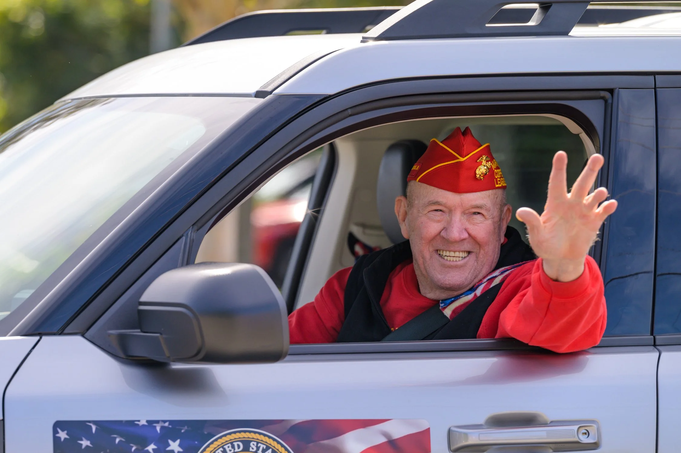 A smiling man in a red veteran's cap and red jacket waving from the driver's side window of a vehicle decorated with an American flag and a badge.