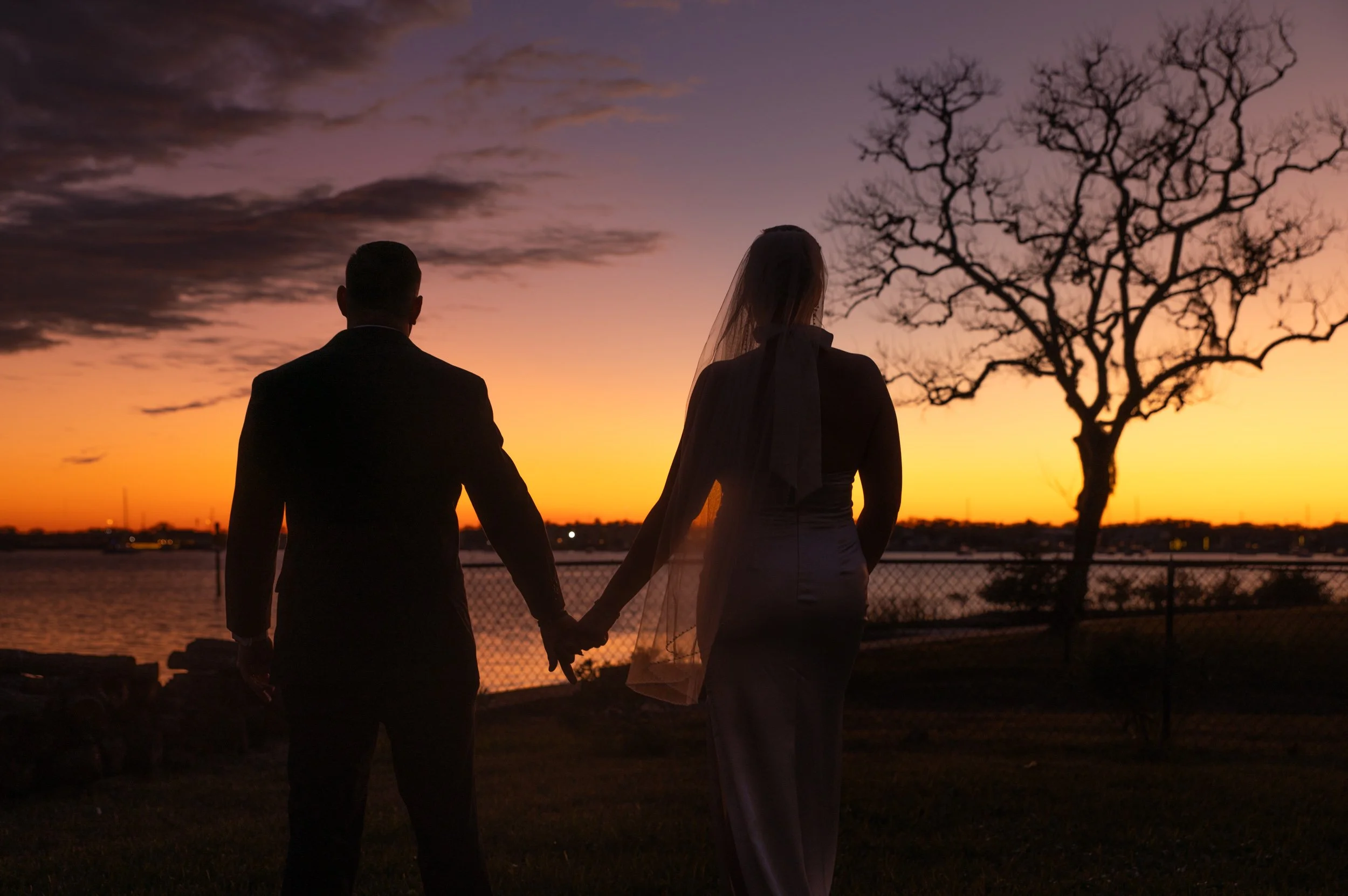 A couple dressed in wedding attire holding hands at sunset by a body of water, with a leafless tree and cloudy sky in the background.