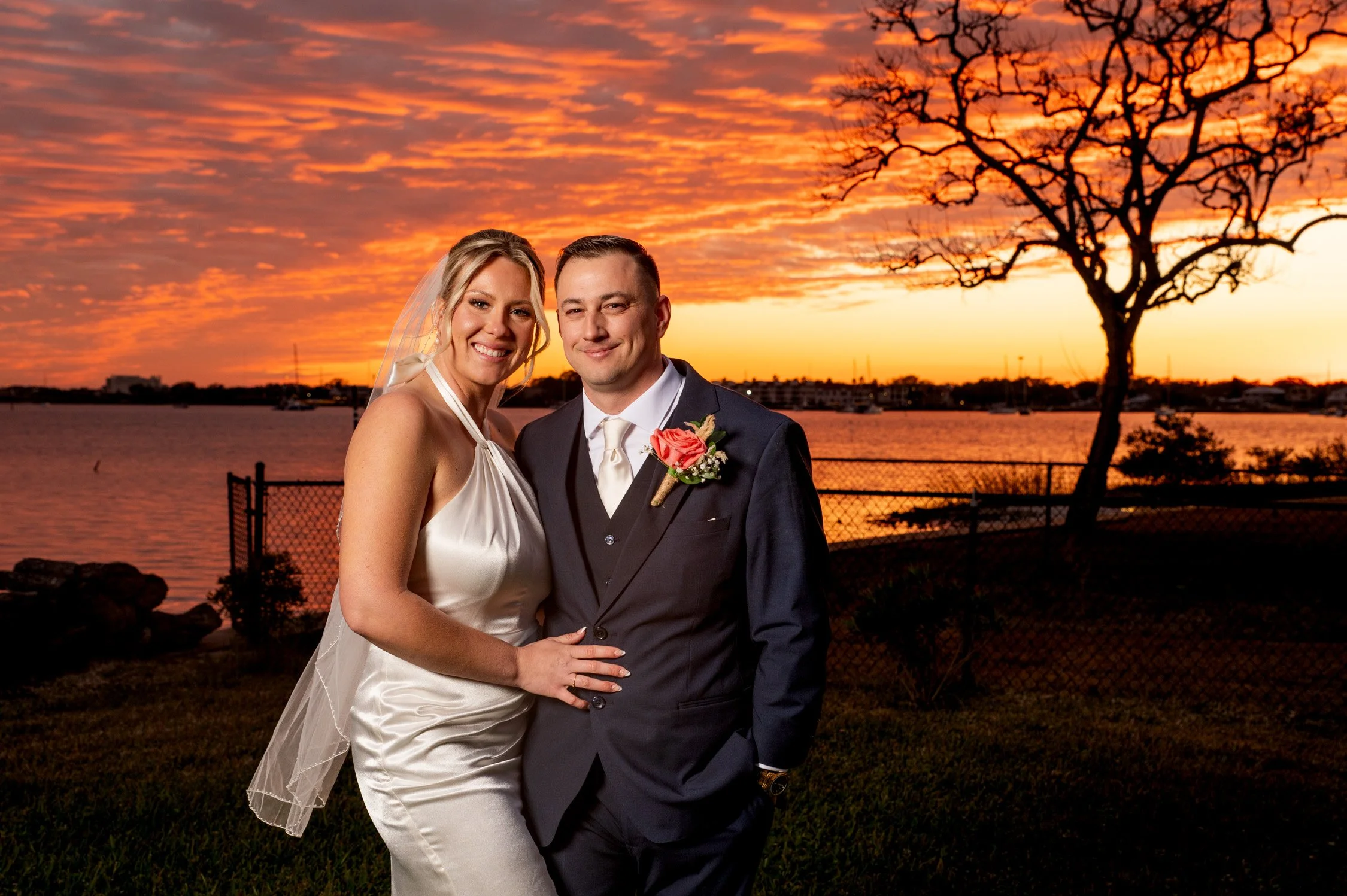 A bride and groom standing together at sunset by a body of water, smiling. The bride is wearing a white satin wedding dress and veil, the groom is in a dark suit with a boutonniere. There is a tree and a fence behind them.