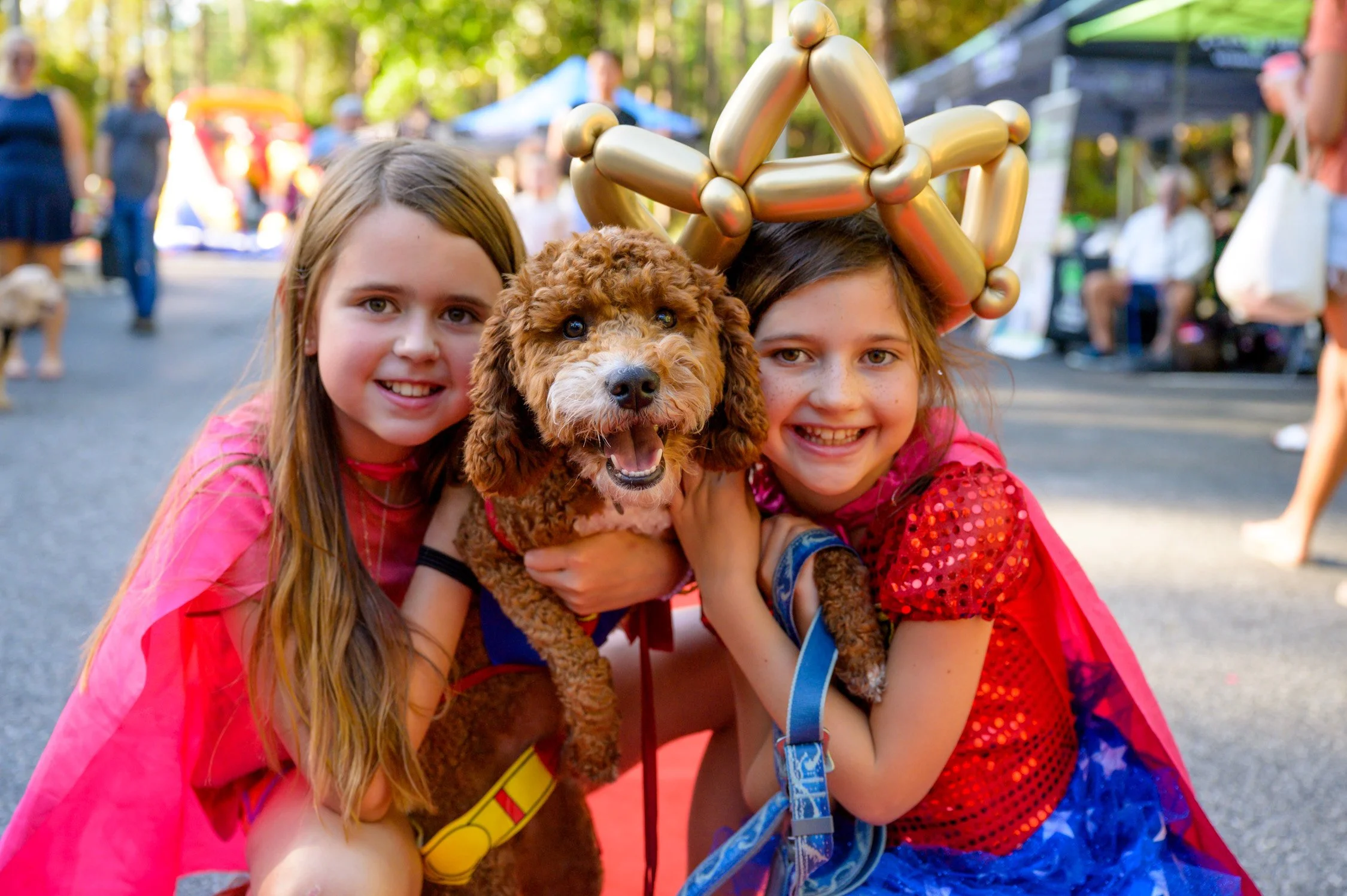 Two smiling young girls in colorful costumes, one wearing a balloon crown, hugging a happy brown poodle dog at an outdoor event with tents and people in the background.