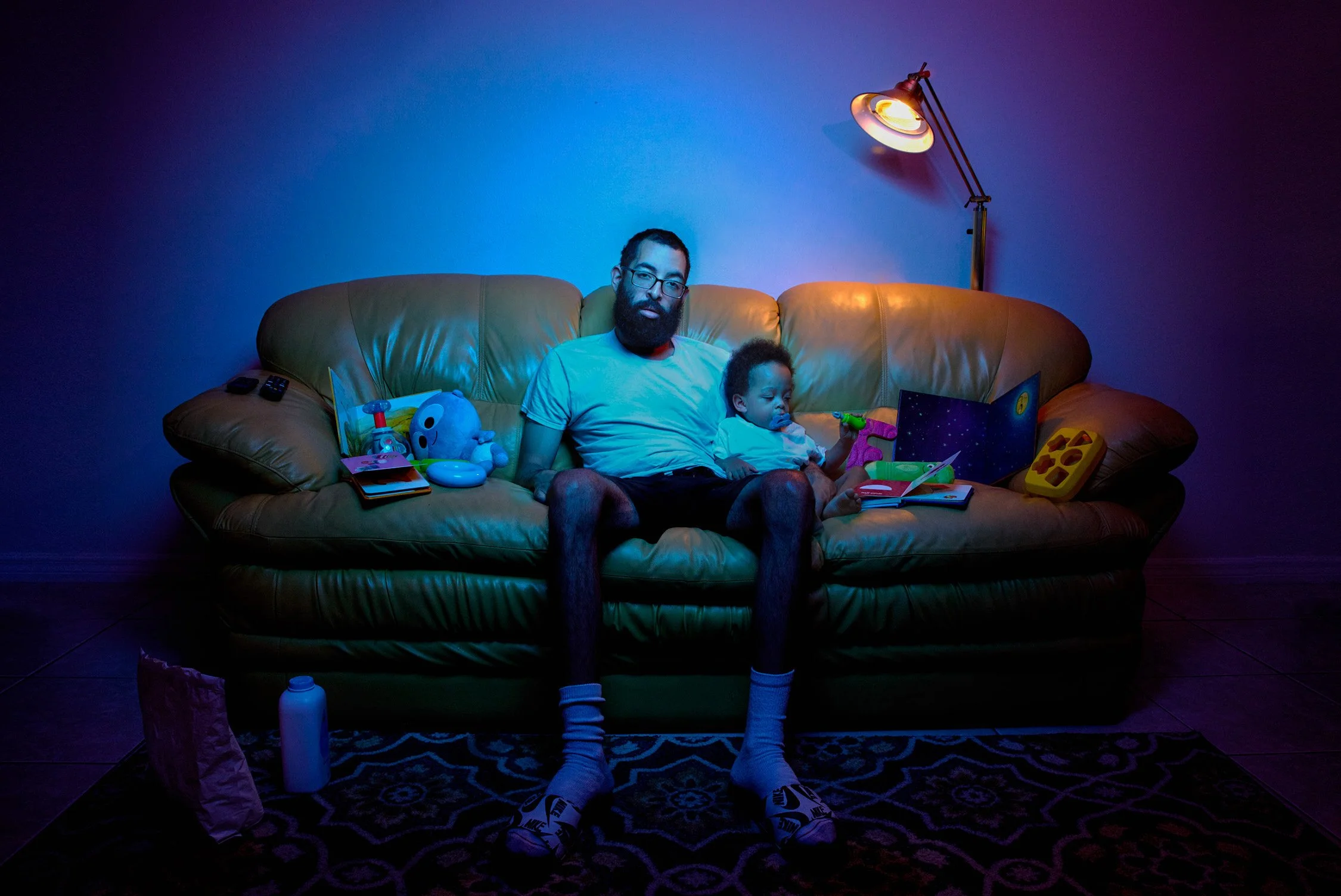 Man and young child sitting on a yellow leather couch, surrounded by toys, in a dimly lit room with a blue and purple glow from a floor lamp.