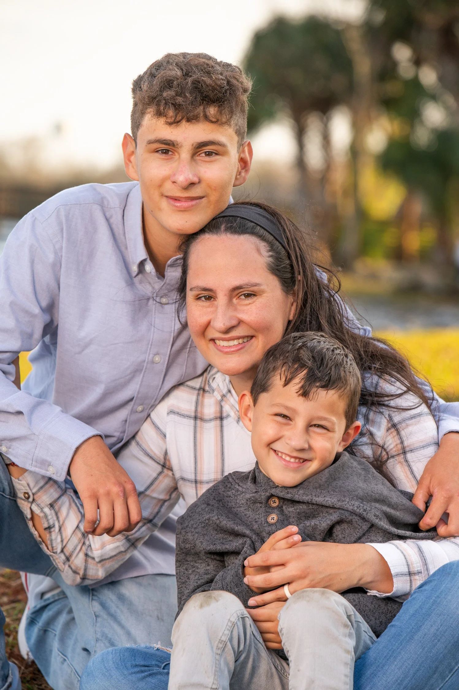 A happy family of four outdoors, sitting on grass, smiling at the camera with trees in the background.