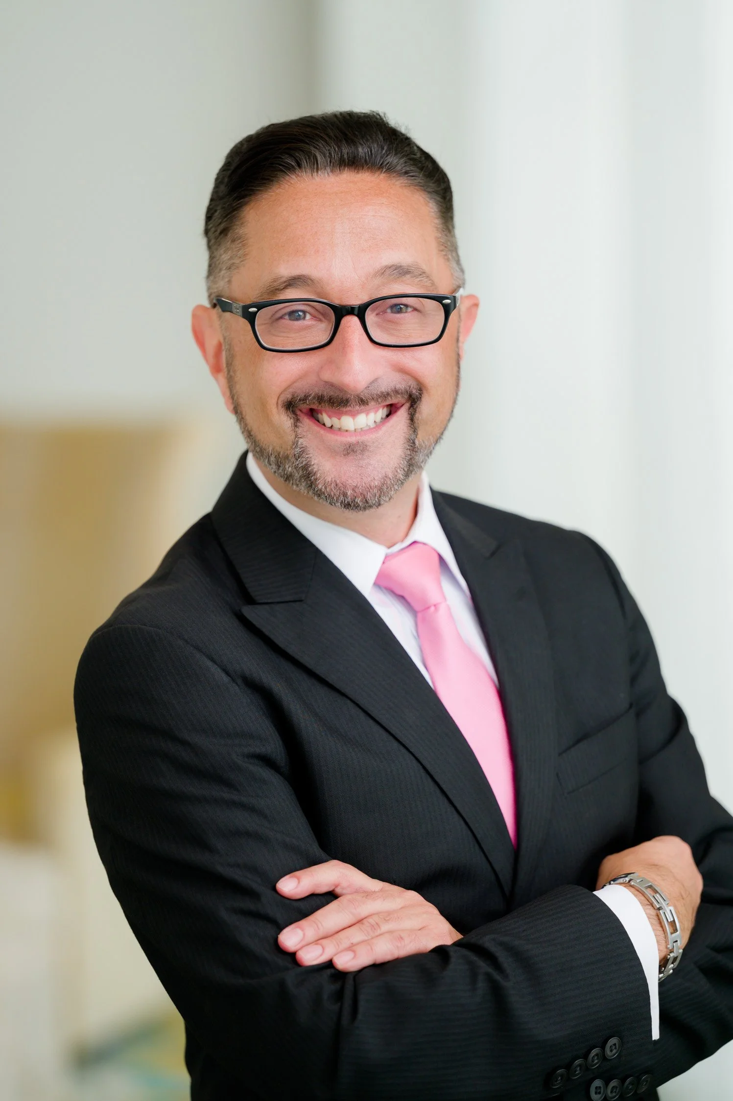 A smiling man wearing glasses, a dark suit, a white shirt, and a pink tie, with arms crossed, in an office setting.