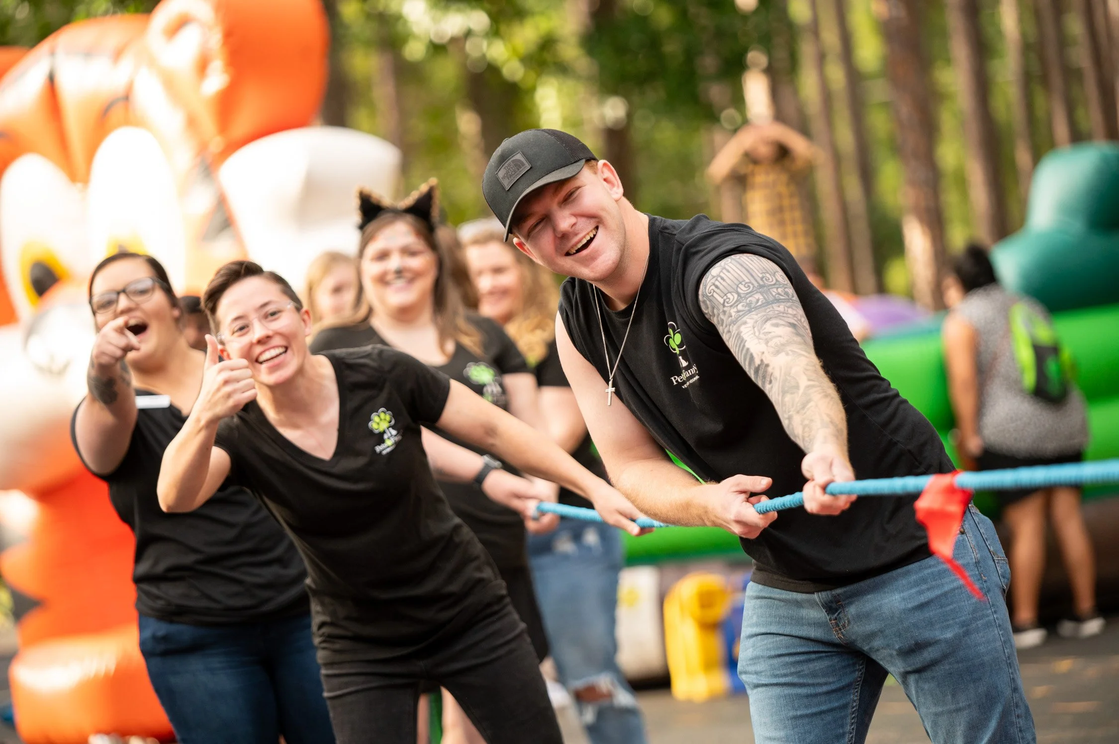 People participating in a tug-of-war game outdoors during a carnival or fair, surrounded by colorful inflatables and trees.
