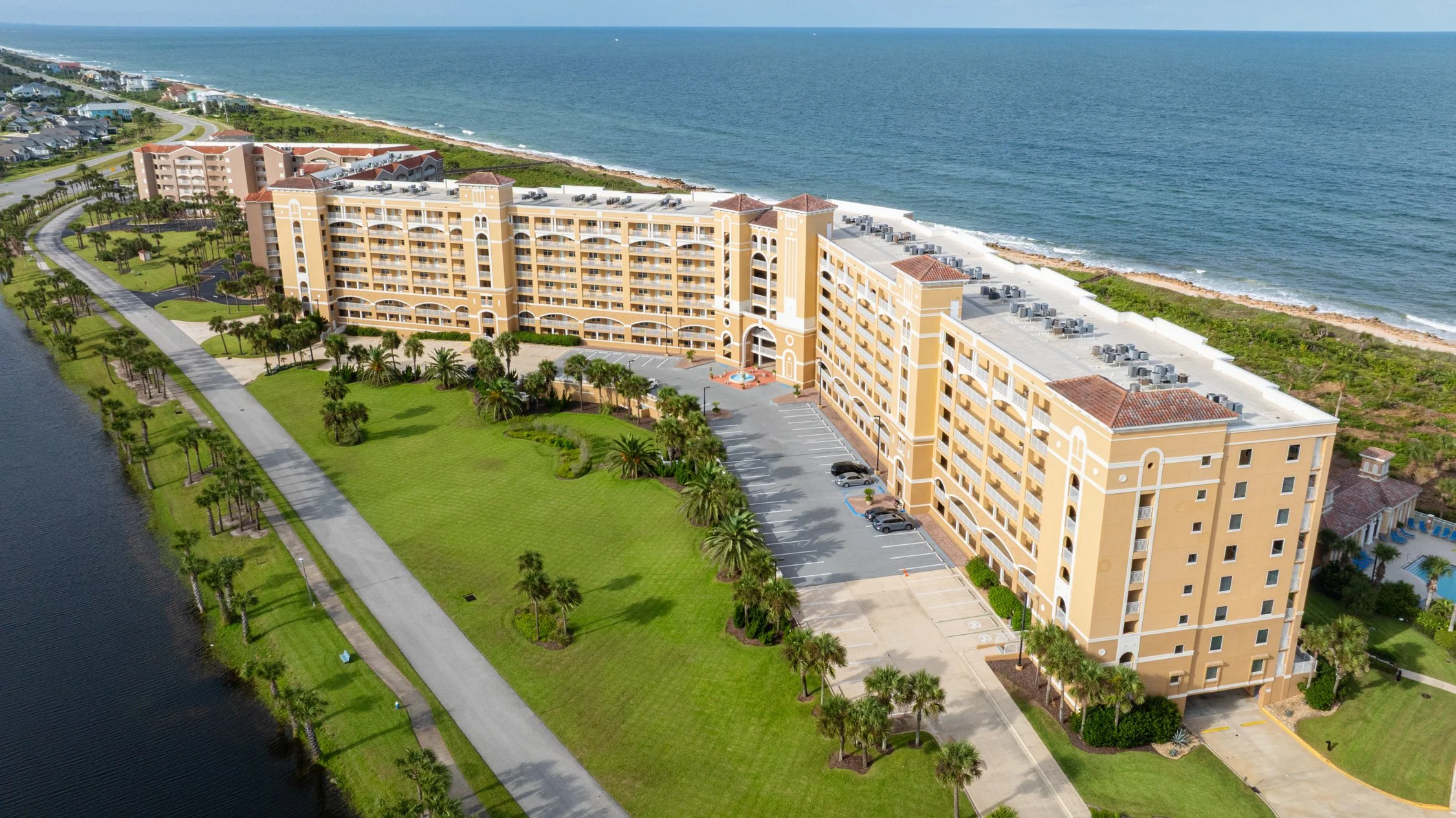 A large beachfront resort with a curved building and a parking lot, surrounded by green lawns and palm trees, overlooking the ocean.