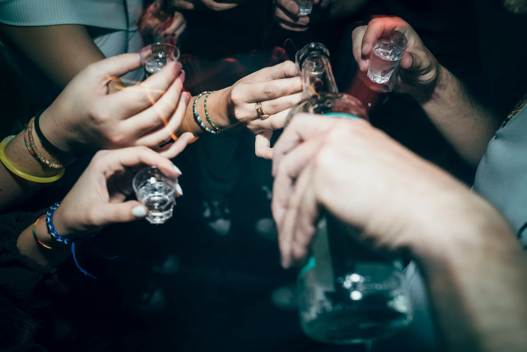 Mehrere Personen stoßen in einer Gruppe mit kleinen Gläsern auf ein gemeinsames Ereignis an, Hände und Armbänder sichtbar.