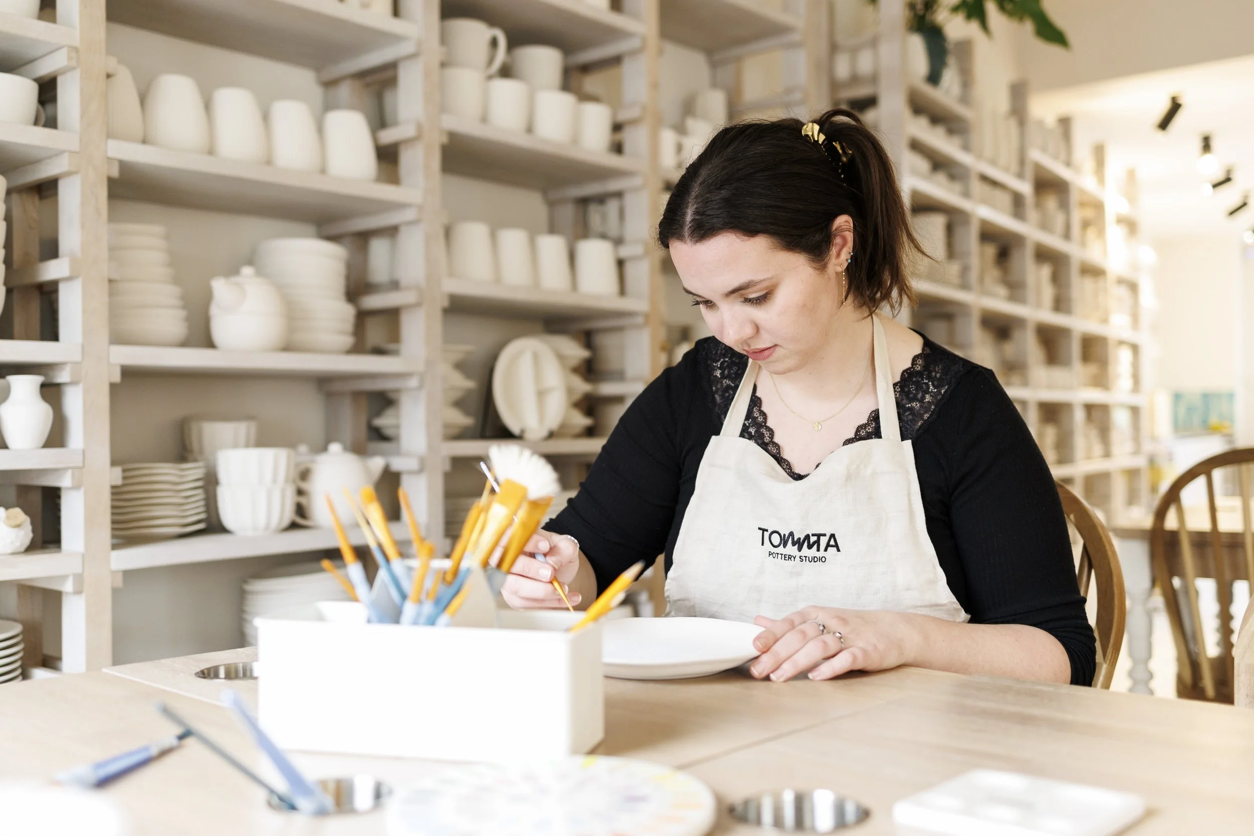 A woman with short dark hair working at a pottery studio, wearing a white apron with the logo 'TOMITA POTTERY STUDIO' on it. She is focused on her craft, with shelves of white ceramic pieces behind her and various paintbrushes and tools in front.