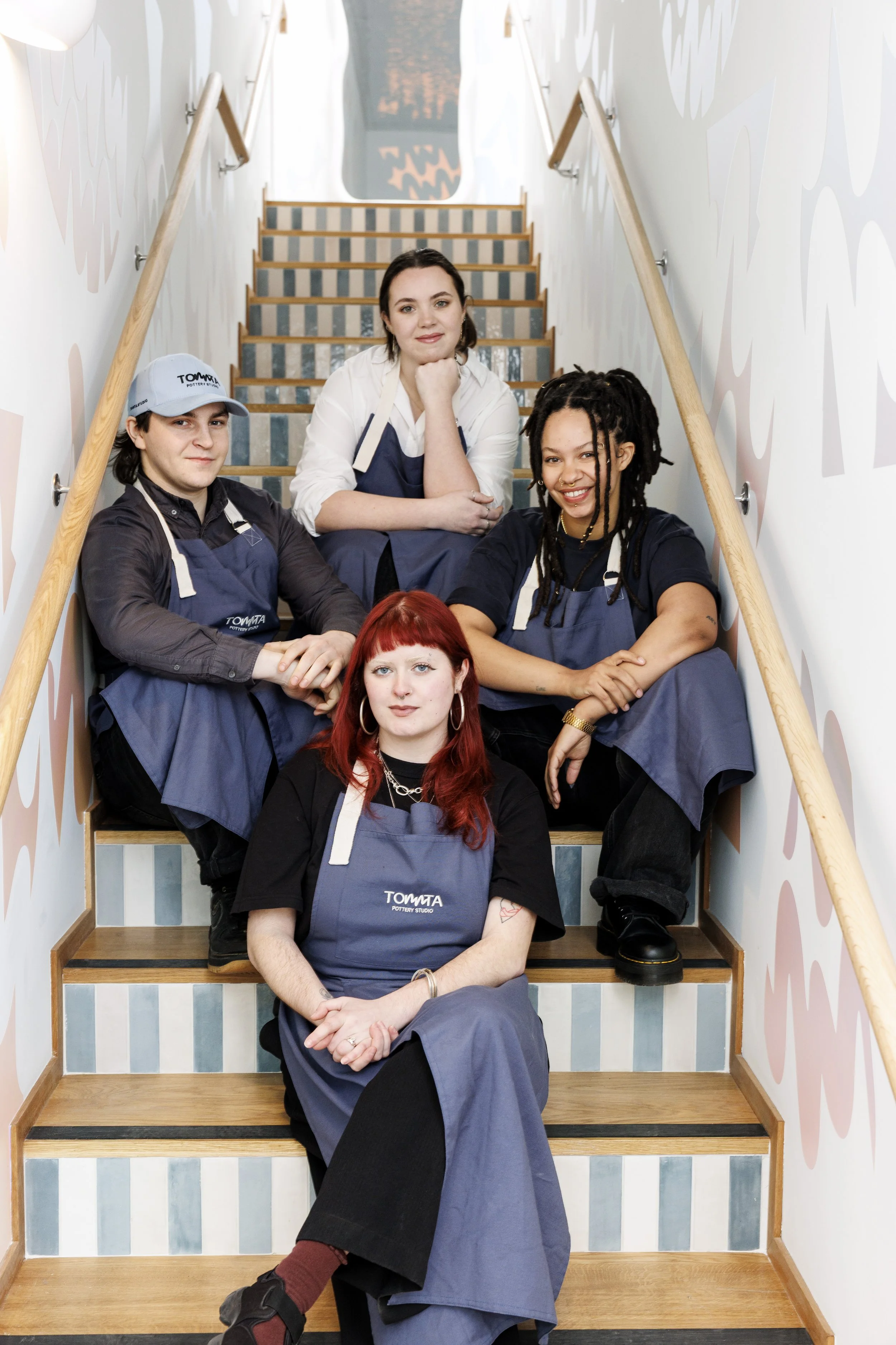 A group of five diverse women sitting on a staircase in a pottery studio, wearing aprons with the logo "TOMITA Pottery Studio," posing for a photo and smiling.