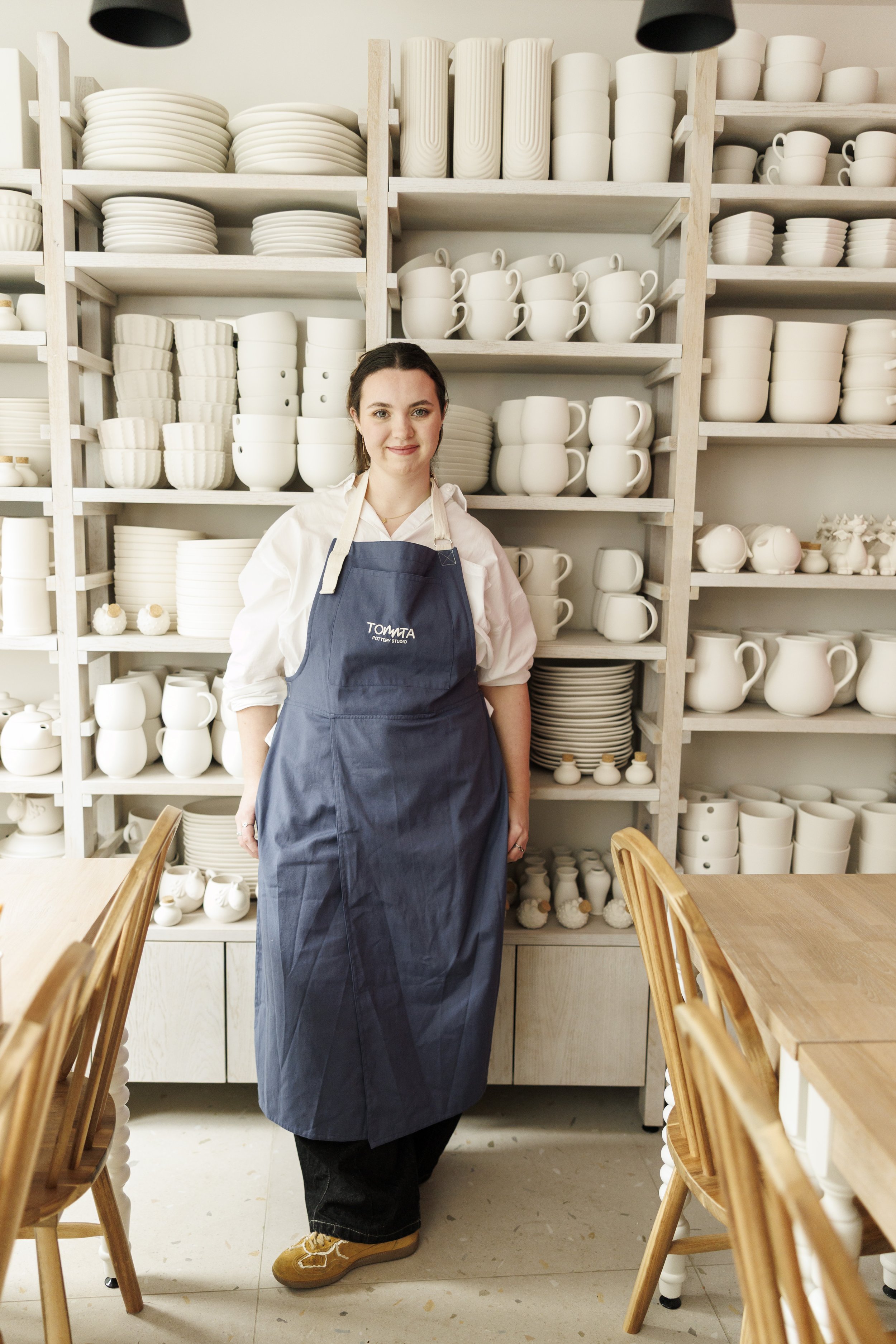 A woman standing in a pottery studio surrounded by shelves filled with white ceramic dishes, cups, and vases, wearing a navy apron with 'TOMA' branding, a white shirt, and brown shoes.