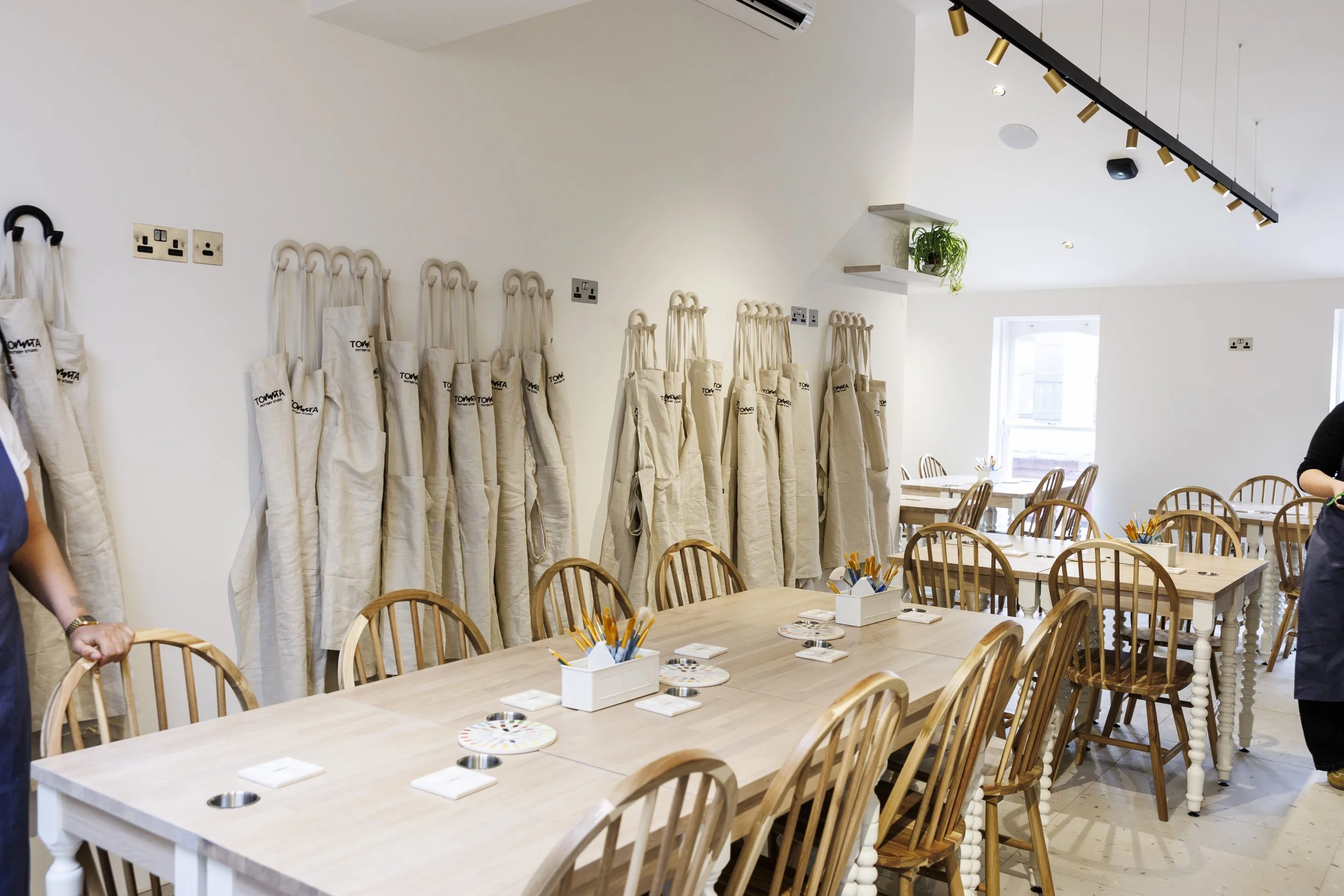 A spacious room set up for a team activity or workshop with multiple beige tote bags hanging on the wall, wooden chairs around long tables with art supplies, and a bright, modern interior with white walls, shelves with plants, and large windows.