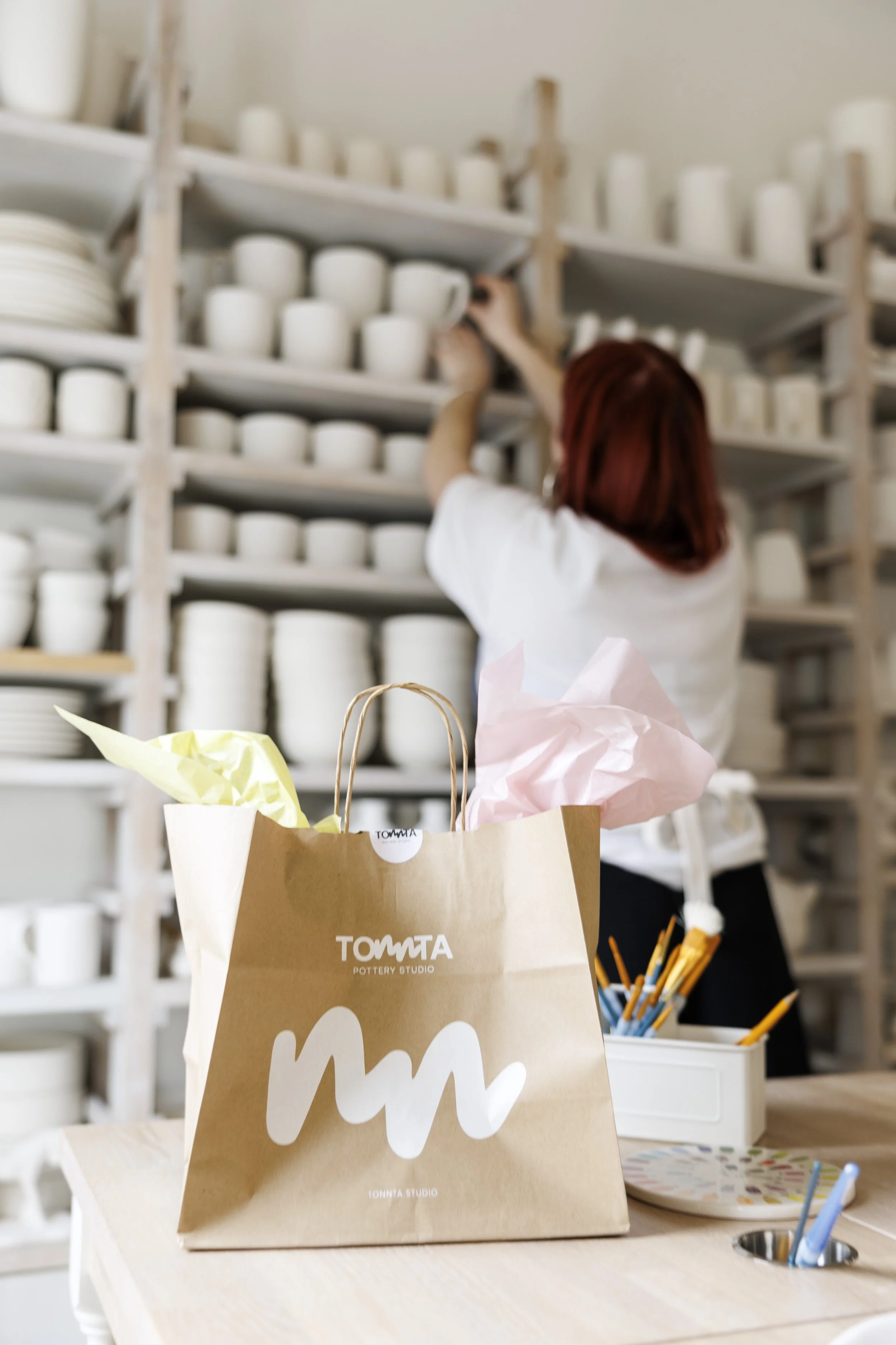 A woman with red hair arranging white pottery on shelves in a pottery studio. There are gift bags and paintbrushes on the table.