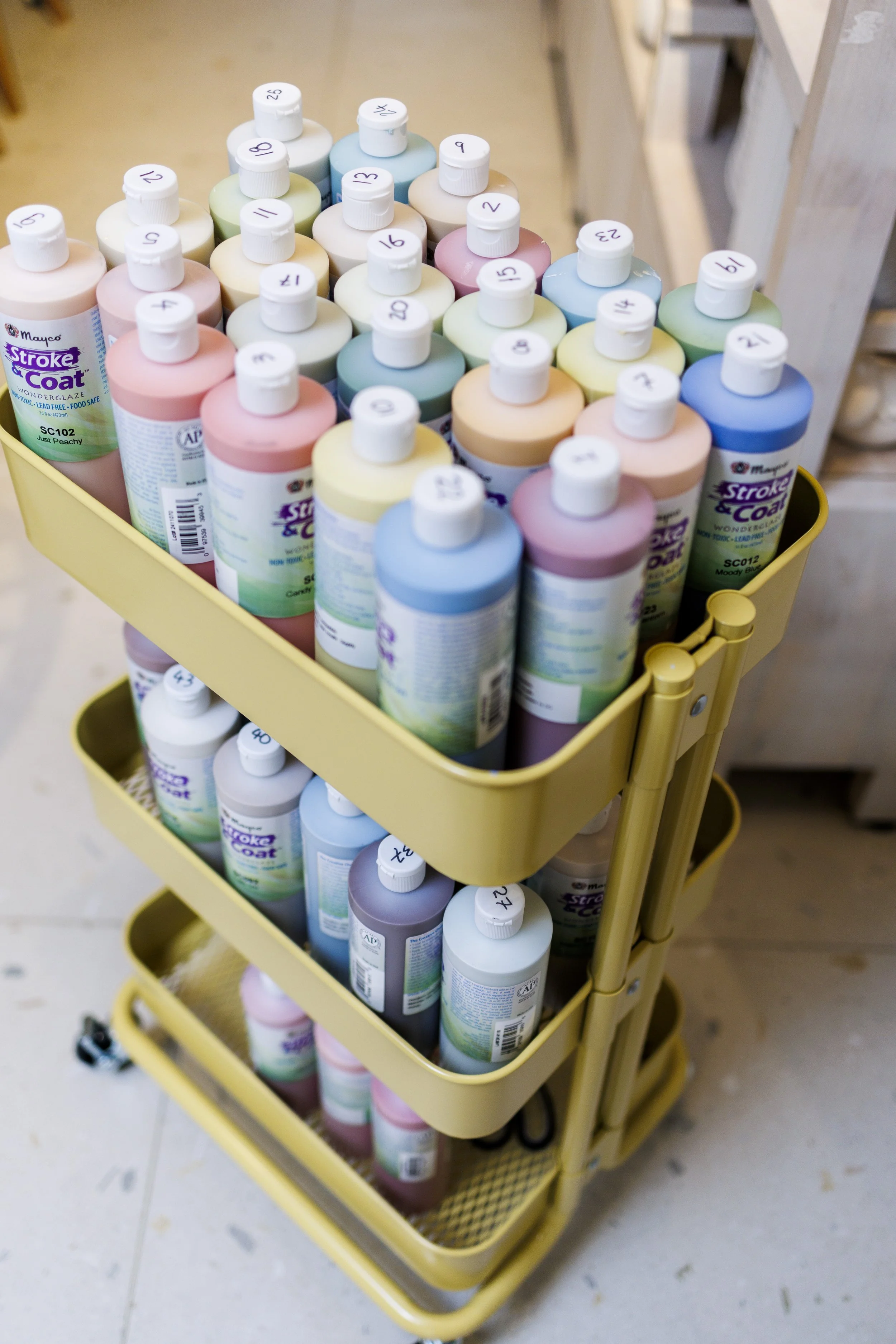A yellow cart filled with bottles of paint, labeled with numbers on the caps, in various pastel colors, with a light-colored floor in the background.