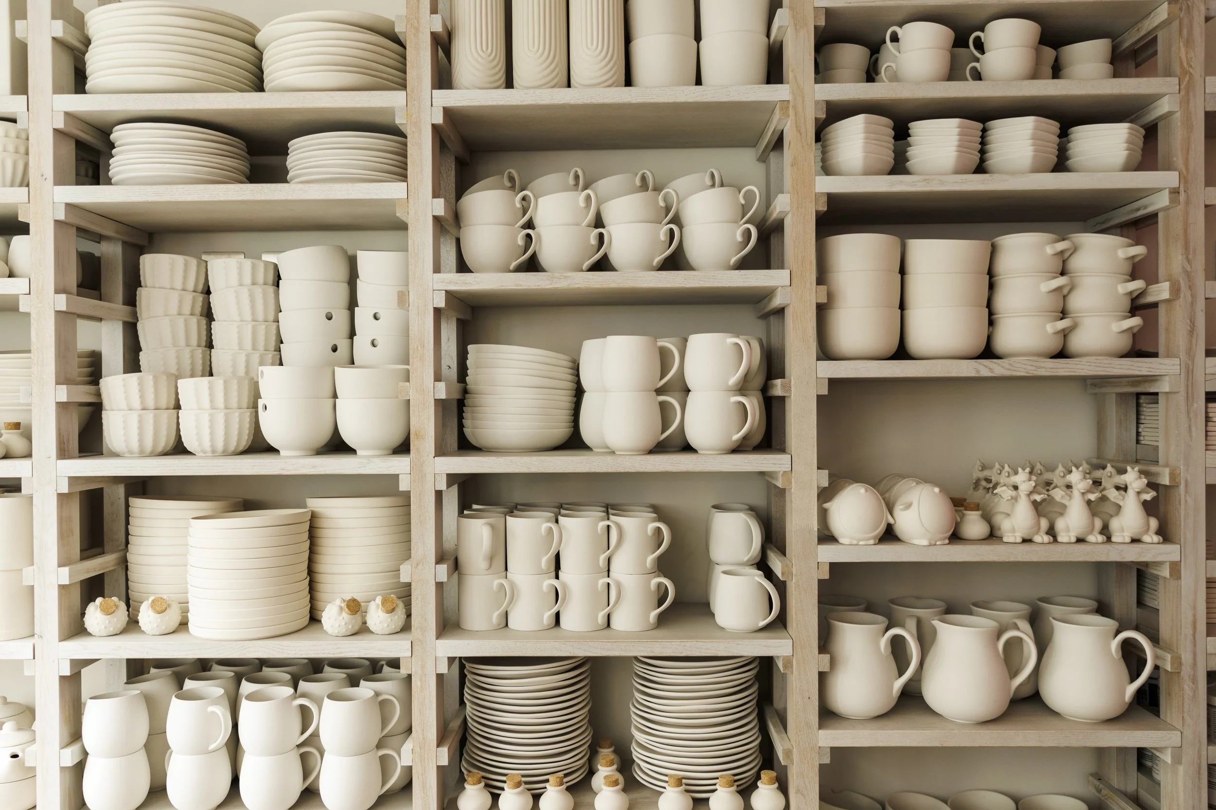 Multiple shelves filled with unglazed ceramic dishware including plates, bowls, cups, mugs, and small decorative items in a pottery studio.